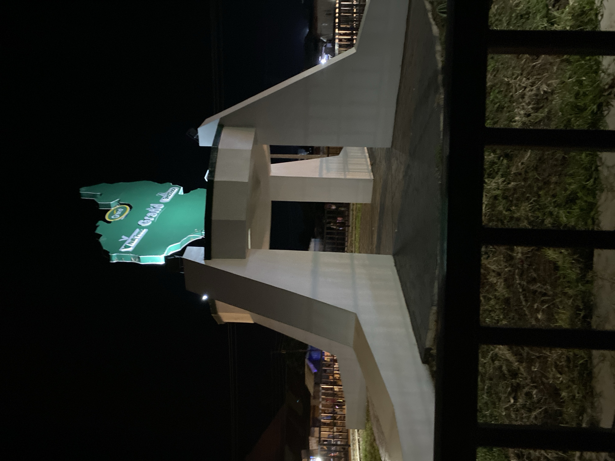 The image depicts a landmark structure at night in Bukuru, Nigeria. The structure is a large, white, arch-like gateway with two angled wings extending outwards from a central opening. Above the opening, a prominent green sign shaped like a map of a region is illuminated. The sign features text that reads "Grand" and other indistinct words, possibly indicating a specific location or area within Bukuru. The sky is dark, suggesting nighttime. In the background, there are blurred lights and structures, hinting at an urban or semi-urban environment. The foreground is partially obscured by a dark railing, with patchy grass and some dried vegetation visible beneath it.