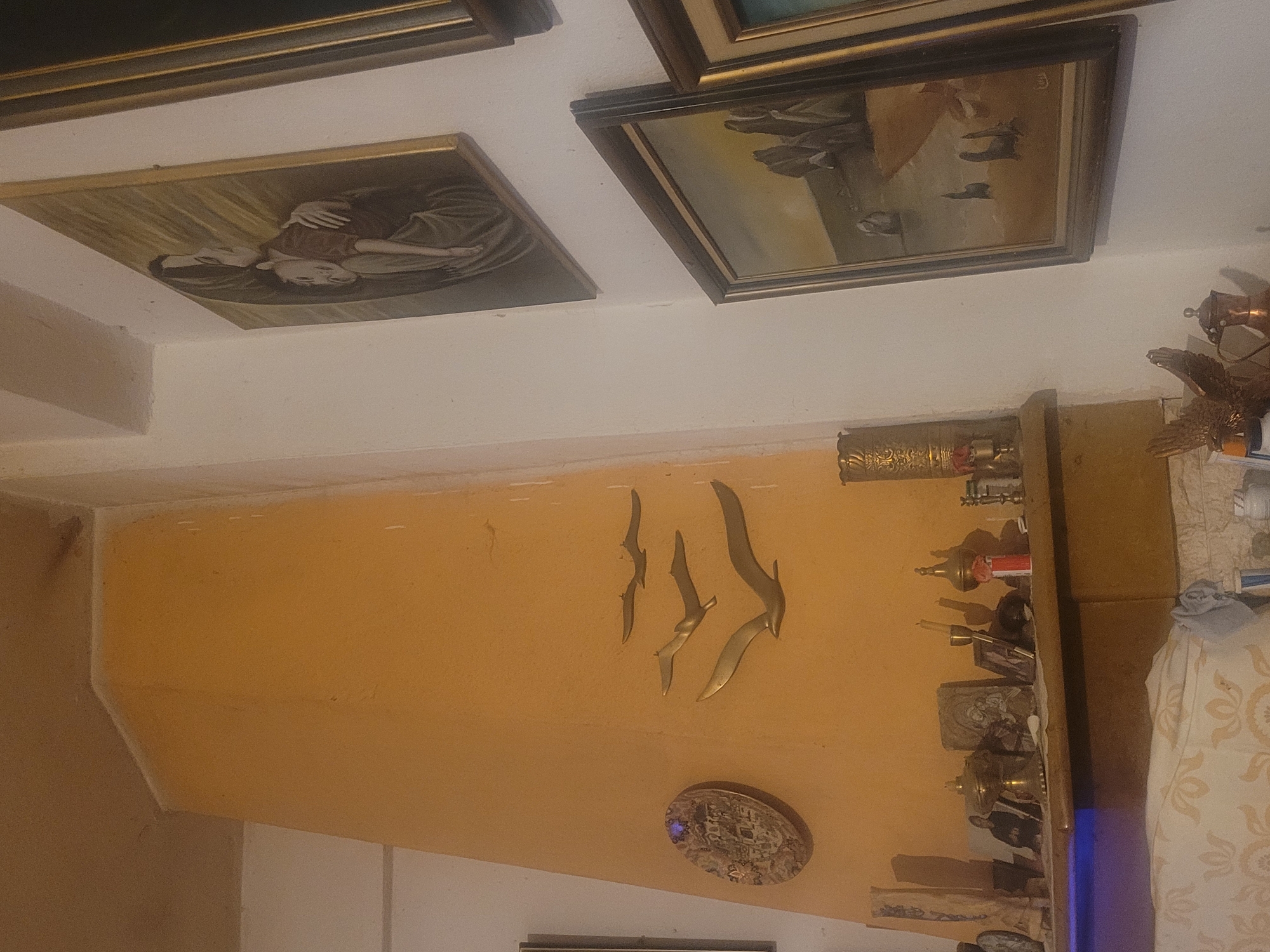 The image displays a room interior, likely a living space, featuring a wall painted in a light orange hue. Attached to the wall are decorative elements including metal bird sculptures and a patterned ceramic plate. A shelf positioned below the birds houses an eclectic collection of items such as brass containers, candlesticks, and decorative figurines. Above, a white ceiling is adorned with three framed paintings. The room's overall decor suggests a personal space with an artistic and collected aesthetic. The exact location of the room is not discernible from the visual information.