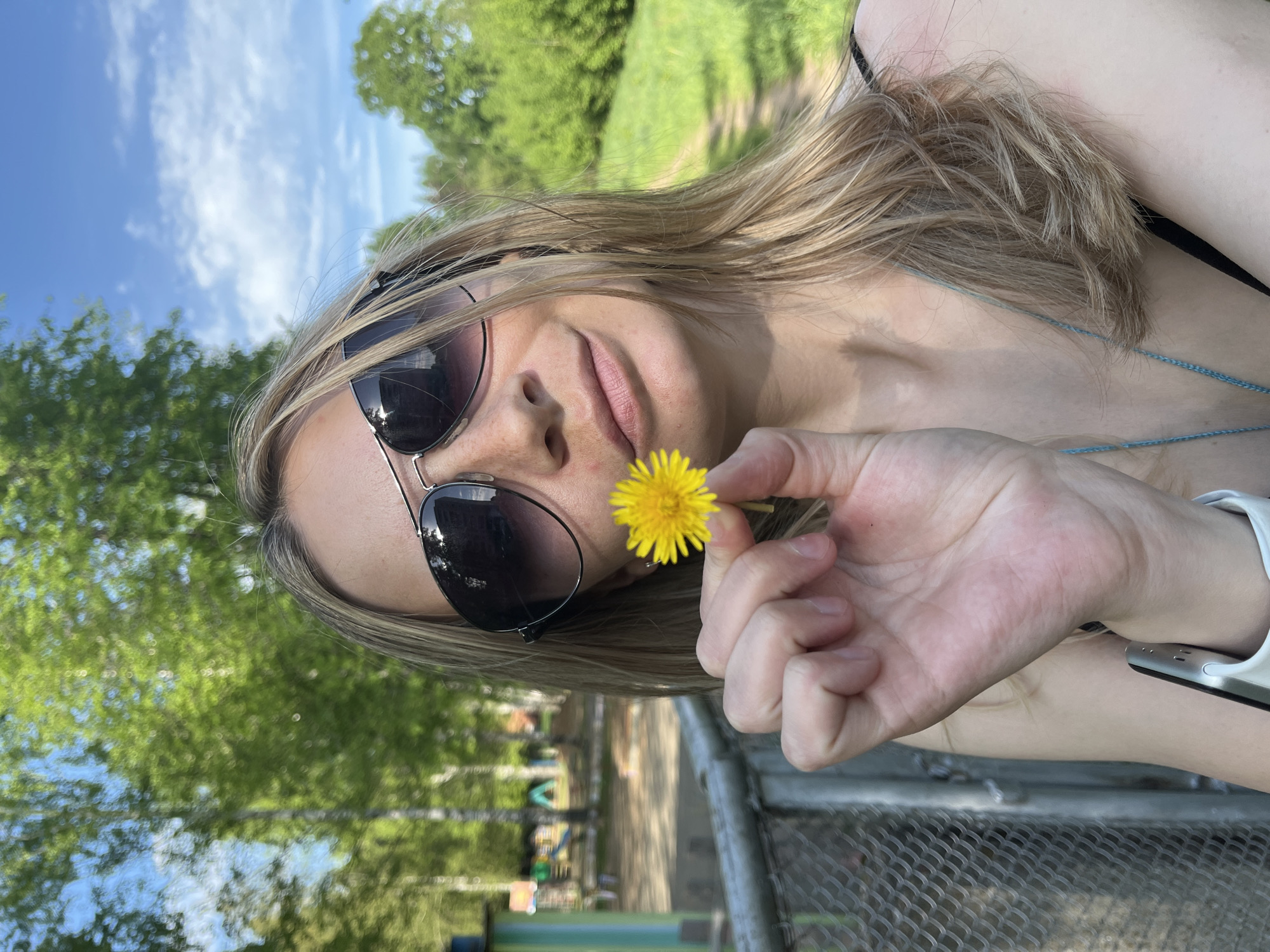 A woman with long blonde hair is holding a yellow dandelion flower in her right hand. She is wearing sunglasses and appears to be outdoors. The background includes trees, a blue sky with clouds, and a chain-link fence. It is likely that this photo was taken in the city of Chaikovsky, Russia, as requested in the prompt. There is a playground structure visible behind the chain-link fence. The woman's wrist is visible with a watch, possibly an Apple watch. She is wearing a black tank top and a necklace.