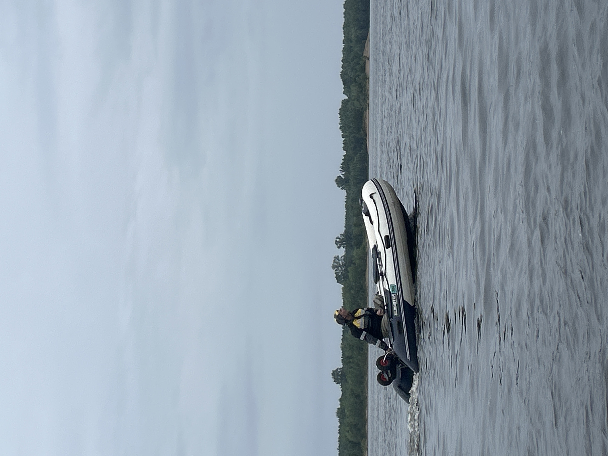 A person wearing a yellow and black life vest is in an inflatable boat on the water. The boat has a blue and white exterior and a dark blue trim. The person is looking out towards the water. There are two small red wheels attached to the back of the boat. In the background, a line of trees forms a dark green barrier along the shore. The sky is overcast and a pale grey. The water appears choppy with ripples and small waves. The image is oriented vertically, with the boat and person on the right side. The overall scene suggests a cool, possibly cloudy day in a natural environment. The text "GLADIATOR" is visible on the side of the boat.