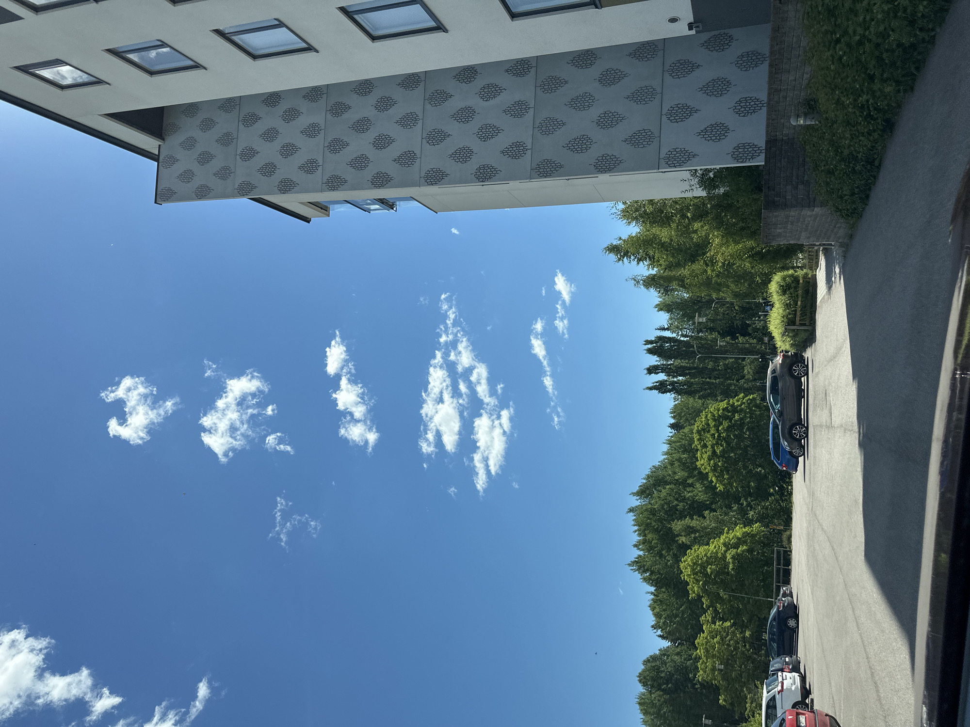 This is a vertical, full-frame outdoor shot on a bright, sunny day. The scene appears to be a residential or commercial area with a modern building on the left and a tree-lined street with parked cars on the right.
The building on the left has a light-colored facade with several square windows, some reflecting the sky. A patterned section of the building features a repeating geometric design. The sky is a vibrant blue with scattered, wispy white clouds.
The right side of the image shows an asphalt road with several cars parked alongside it. The cars are of various colors, including gray, blue, black, white, and red. Lush green trees line the street behind the parked cars, suggesting a pleasant, possibly summer, season. A hedge is also visible along the edge of the property.
The lighting indicates it is daytime, likely mid-morning to early afternoon, given the clear skies and strong sunlight. There is no discernible activity or event taking place; the image captures a static view of the environment. No people are visible, and there is no visible text in the image. The overall mood is peaceful and serene, with the bright weather contributing to a feeling of openness and clarity.