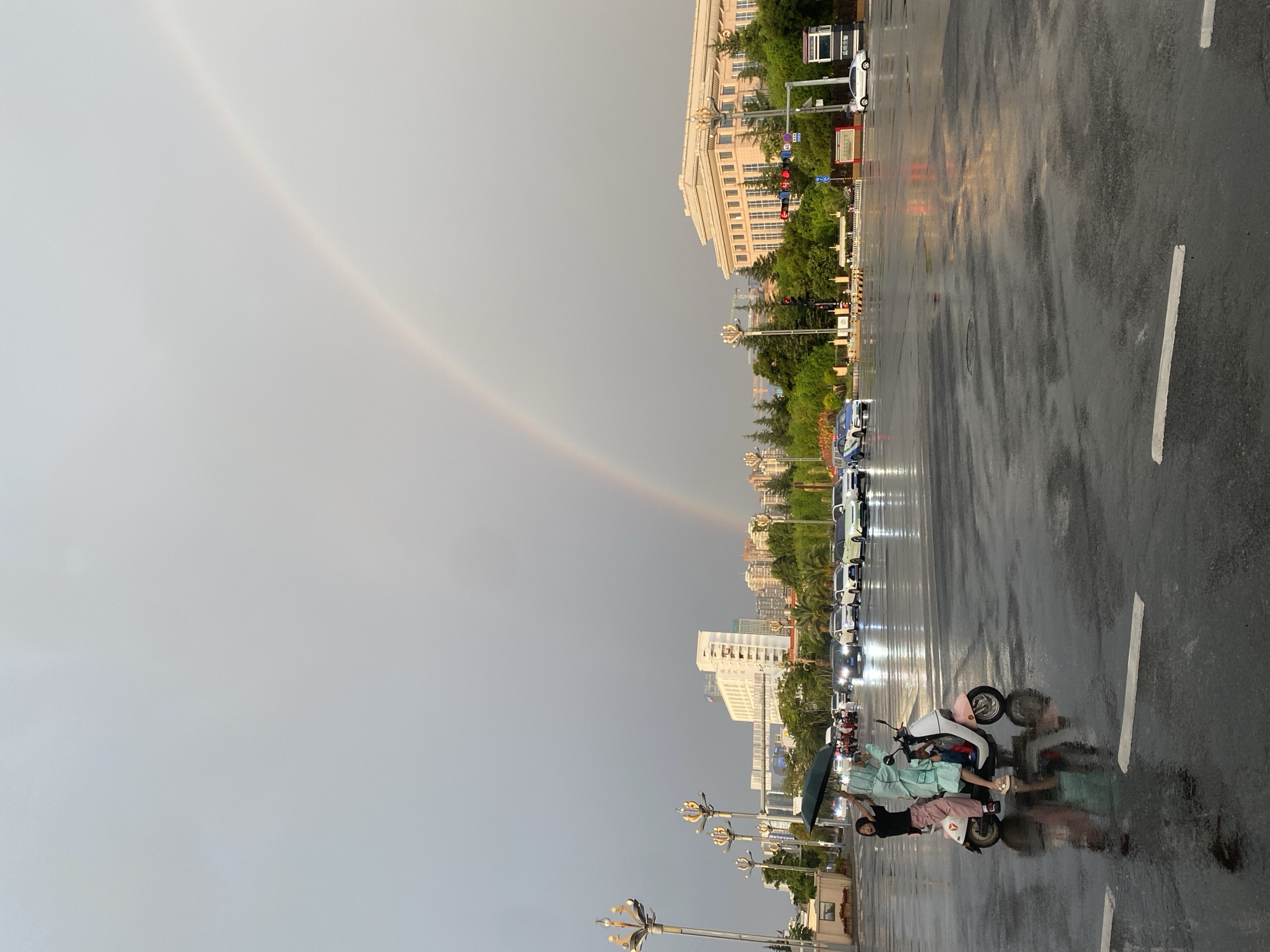 In Xishan District, China, a street scene unfolds under a grey, overcast sky, bisected by a faint rainbow arching across the upper portion of the frame. The wet asphalt reflects the muted light, indicating recent rainfall. In the foreground, two individuals are seated on a pink and white scooter. The person in the rear, wearing a light blue rain poncho, holds a black umbrella aloft. The person in front, dressed in a black shirt and pink pants, leans forward, seemingly looking towards the left. Their reflections are visible on the wet road surface.
The background features a city environment with various buildings and infrastructure. To the left, a row of ornamental streetlights stands prominently. Beyond them, a multi-story white building with numerous windows is visible, alongside other urban structures and greenery. To the right, a large, light-colored building with classical architectural elements dominates the mid-ground. Traffic is present, with several cars, some of which are illuminated by their headlights, moving along the wet road. A white car is parked on the right side of the image. The overall atmosphere is one of a transitional moment, with the remnants of rain and the appearance of a rainbow suggesting a clearing sky.