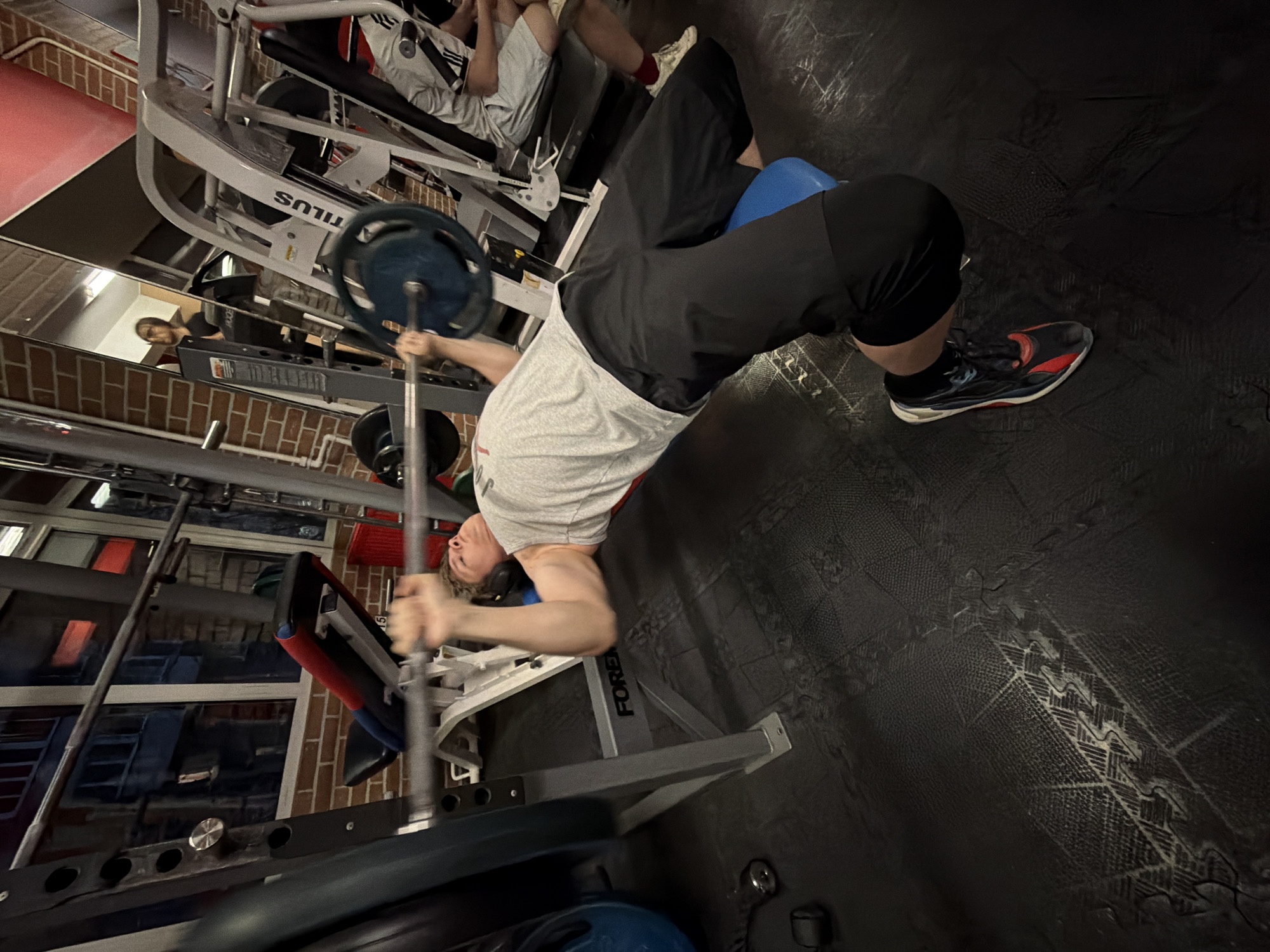 A man is performing a barbell hip thrust exercise at a gym. He is wearing a gray t-shirt and black shorts. A blue pad is placed under his upper back for comfort. The barbell is loaded with weight plates. He is lying on his back, with his knees bent and feet flat on the floor. The gym has various weight machines and equipment. There are other people present in the gym. The image location details requested cannot be identified.