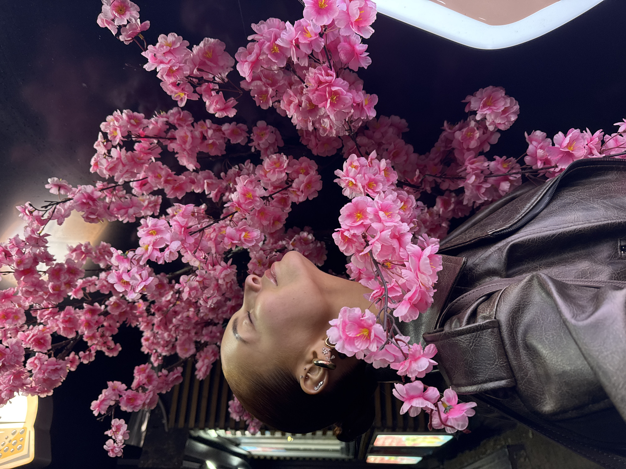 A person is captured in profile, looking upwards with a serene expression, as if observing or contemplating the scene above. They are positioned beneath a dense canopy of bright pink, artificial cherry blossom-like flowers, which dominate the upper portion of the image. The person's head is tilted back, with eyes closed or downcast. They wear a brown, textured jacket, possibly leather or faux leather. The left ear is adorned with several gold-toned earrings, including a prominent hoop, small studs, and a decorative ear cuff.
The setting appears to be an interior space or a covered outdoor area, possibly a themed restaurant or bar in Izhevsk, Russia, given the artificial nature of the flowers and the dark background. Some ambient light sources, a soft white glow and a yellowish fixture, are visible at the edges of the frame, suggesting a nighttime or dimly lit environment. The profusion of pink flowers, with their uniform petals and yellow centers, creates a vibrant contrast against the dark background. There is no visible text in the image.