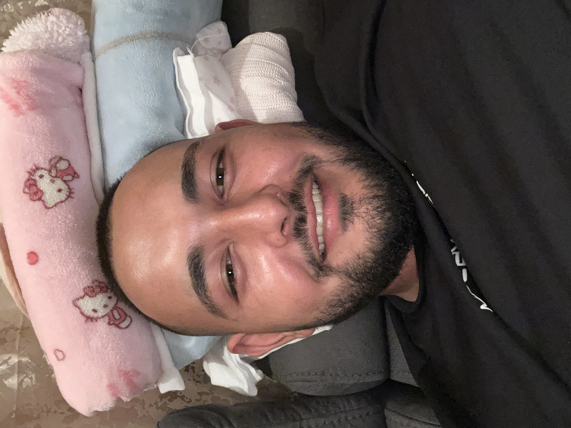 A man with a beard is reclining, his head resting on a pink Hello Kitty blanket and other soft items. He is smiling and looking towards the camera. He is wearing a dark shirt. The location appears to be indoors. There is no clear indication this picture was taken in Razhevo Konare, Bulgaria.