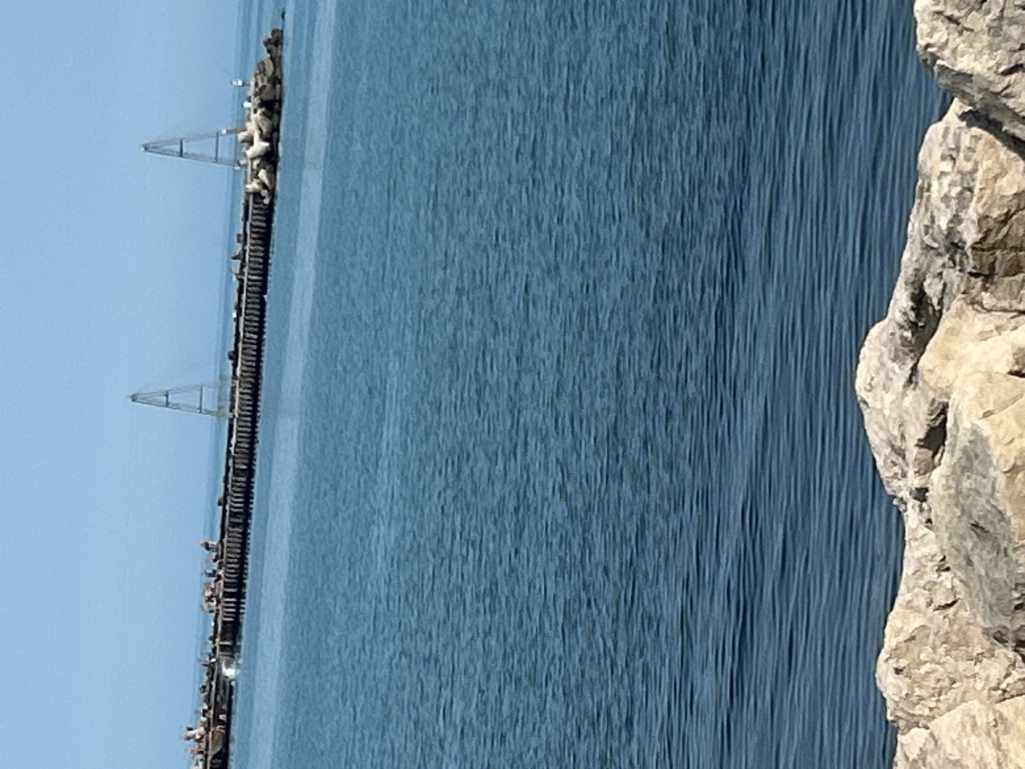 The image displays an outdoor, daytime scene of a body of water and a pier or breakwater under a clear sky. A vast expanse of blue-green water, characterized by subtle ripples, dominates the mid-ground. Extending horizontally across the mid-distance is a long, dark-colored pier or breakwater structure. Multiple small human figures are visible along the pier, particularly clustered on the left side, suggesting activity such as walking or congregating. One individual is discernible on the far right end of the pier, appearing to be engaged in fishing. Two prominent, light-colored, A-frame-like triangular structures, possibly navigational aids, stand erect on the pier at distinct points. The pier's rightmost section transitions into a fortified mass of large, light-colored rocks, likely riprap or tetrapods, on which a small white sign or marker is mounted. The upper portion of the image shows a clear, light blue sky. The foreground contains a collection of large, irregularly shaped, light-colored rocks, forming a textured embankment along the water's edge. The location is in the city of Unknown, Unknown.
