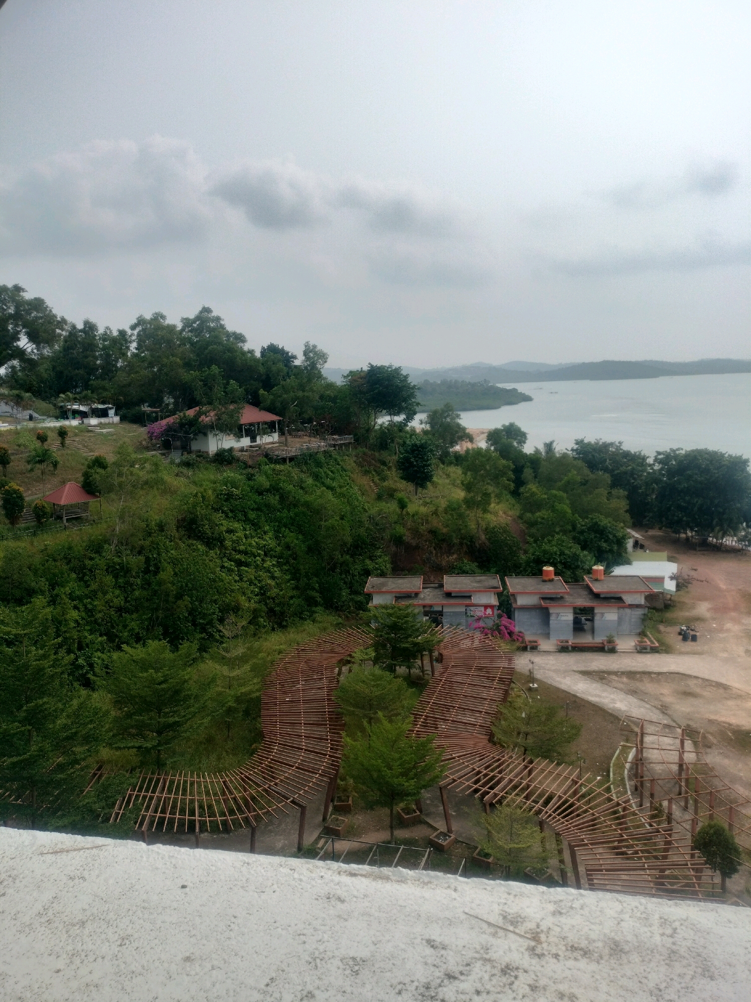 The image depicts an elevated, exterior view of a park-like setting in Batam, Indonesia. In the foreground, a white, textured surface, possibly a wall or railing, dominates the bottom portion of the frame. Beyond this, a winding wooden structure, resembling a pathway or canopy framework, curves through lush green vegetation.
Further back and to the left, a cluster of buildings is visible on a hillside. These include a white building with a red roof, a smaller red-roofed gazebo, and other structures integrated into the landscape. The buildings are surrounded by dense foliage and trees.
To the right of the main buildings, a row of modern, low-lying structures with grey walls and red roofs is situated. These appear to be part of a facility, with visible benches outside. Dirt paths and some scattered trash bins are also present in this area.
The background reveals a body of water, likely the sea, stretching towards a distant horizon where faint outlines of hills and landmasses can be seen under an overcast sky. The overall impression is that of a scenic overlook with recreational facilities, possibly part of a tourist attraction or a public park in a coastal area of Batam. No people are clearly visible in the image.