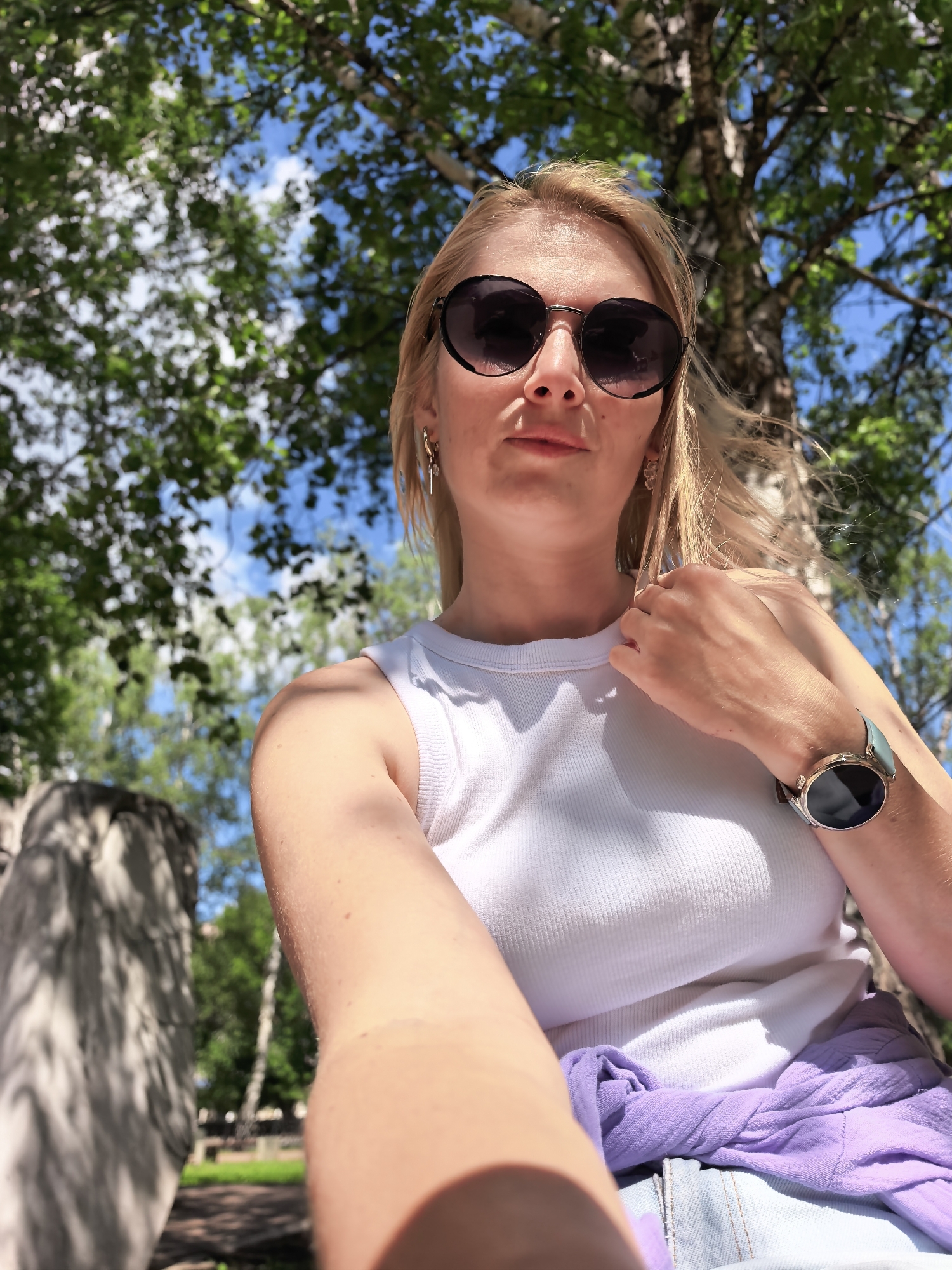 In this selfie, a blonde woman wearing a white tank top and sunglasses is captured outdoors, with trees and blue sky visible in the background. A light purple garment is draped around her waist, and she wears a watch on her left wrist. The image appears to be taken during daylight. The location is not specified in the image or accompanying data.