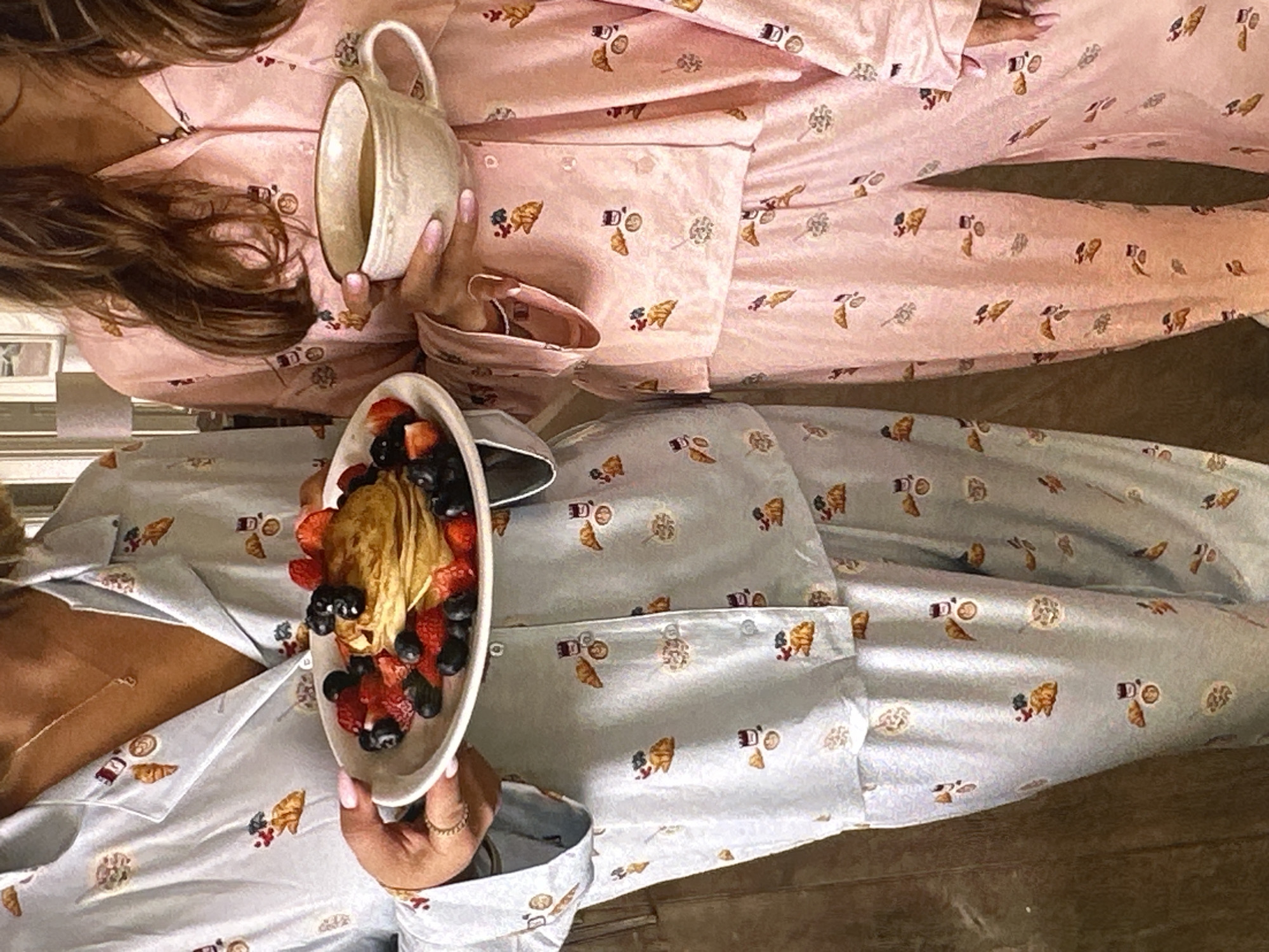Two people are lying parallel to each other, wearing matching pajama sets with a food-themed pattern. The pajama sets are different colors: one is pink and the other is light blue. The person in the pink pajamas is holding a cream-colored mug. The person in the light blue pajamas is holding a white oval plate with a stack of pancakes topped with strawberries, blueberries, and blackberries. The background appears to be a floor, possibly wood, and the setting implies an indoor location. There is no indication that this image was taken in Yerevan, Armenia.