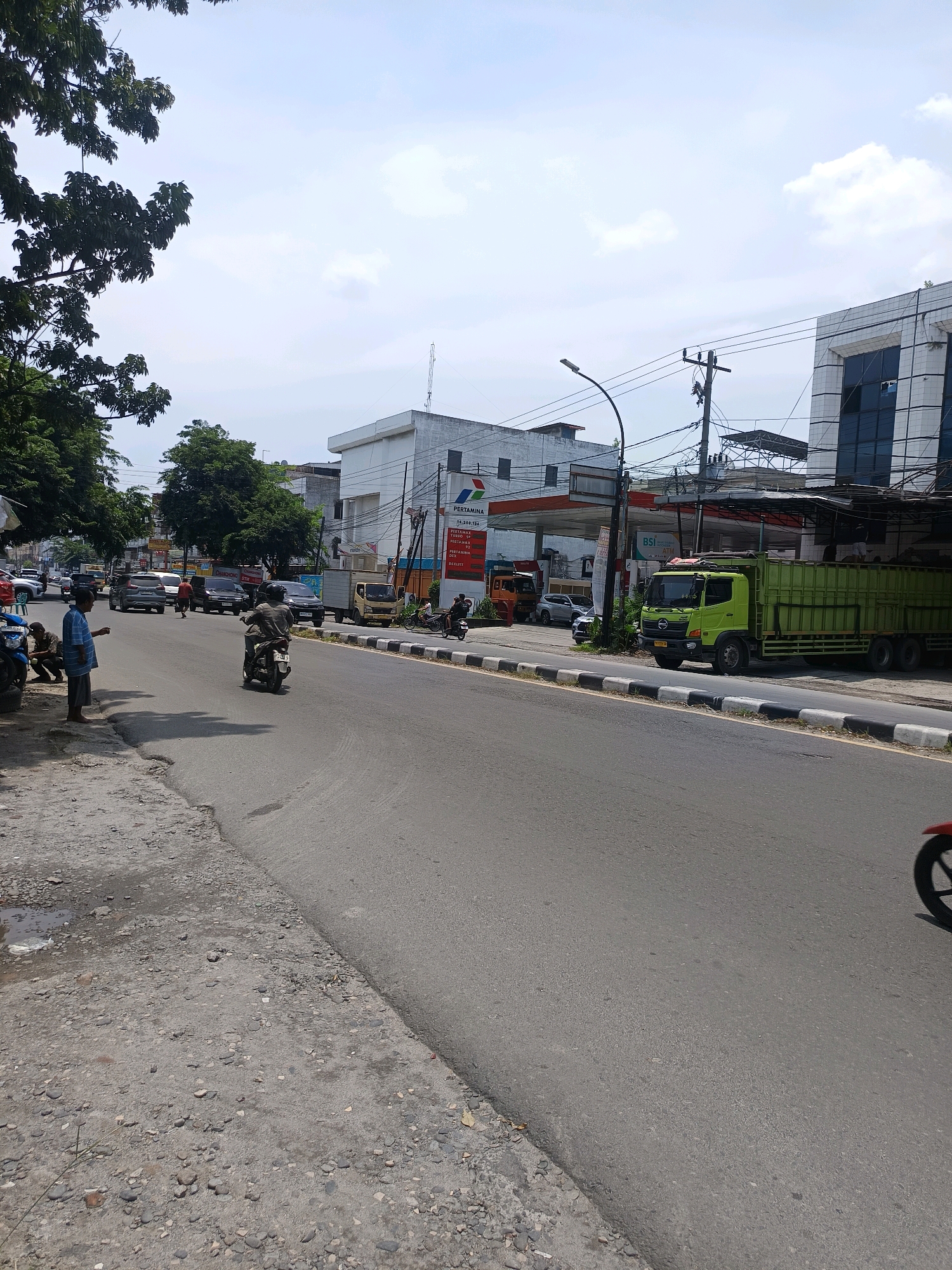 This is a street scene in Percut Sei Tuan, Indonesia, on a clear day with scattered clouds. The image captures a road with various vehicles, buildings, and people. On the right side of the road, a green flatbed truck is parked, and beyond it, a modern building with dark windows and a white facade is visible. Further down the street, a Pertamina gas station is present with visible pricing information displayed. A motorcycle is in motion in the foreground, heading away from the camera. On the left side of the road, a person in a blue and white striped shirt stands on the side of the road, and two individuals are seated nearby. Trees line the left side of the road, casting shadows. The road itself is paved, with a distinct curb and a strip of black and white markings separating the traffic lanes. The environment appears to be a commercial or mixed-use area within the city.