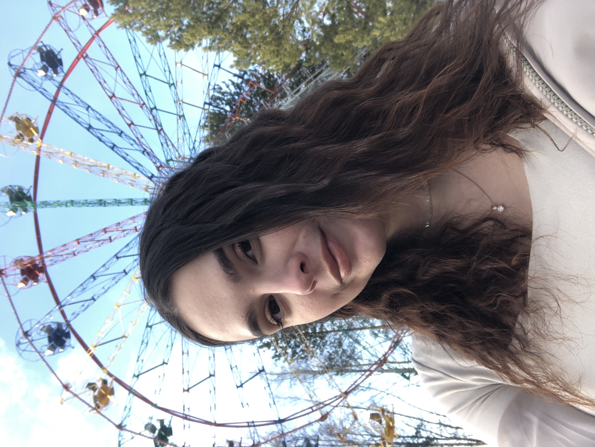 A young female individual is depicted in a close-up, outdoors selfie or portrait, looking towards the camera with a slight smile. She possesses shoulder-length, wavy, dark brown hair and a fair complexion. She wears a light-colored (cream or light beige) top, a subtle choker, and a necklace featuring a small, clear pendant. The background prominently displays a large Ferris wheel with a distinctive red and blue metallic framework, equipped with various multicolored gondolas (yellow, blue, green, red) against a clear light blue sky with sparse white clouds. To the right, green coniferous trees are visible, while to the left, bare deciduous trees are present. This scene is situated in the City Park of Chaikovsky, Russia, where the Ferris wheel is a notable feature.