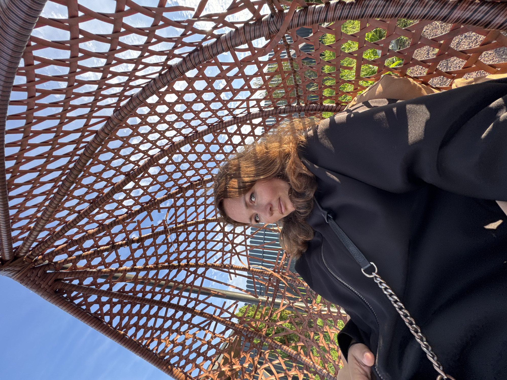 A woman with auburn hair is seated in a brown rattan hanging chair outdoors. She is wearing a black hooded jacket with a silver zipper and a black handbag with a silver chain strap. The chair is suspended from a metal pole and has an intricate woven pattern. Through the open weave of the chair, a wooden fence, green grass, and a gravel path are visible. The sky above is blue with scattered white clouds. The scene appears to be in Kempelevo, Russia, given the information provided.