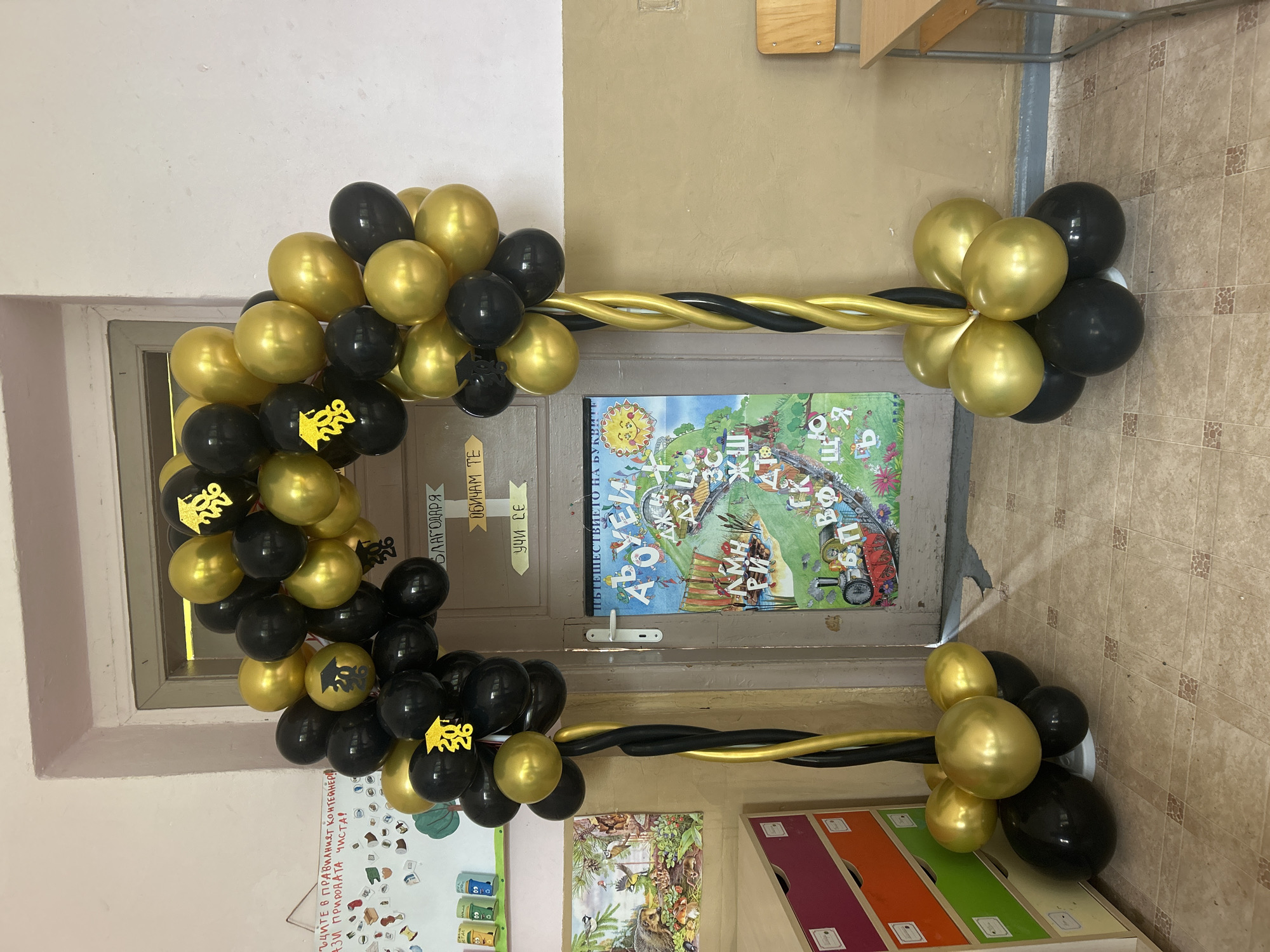 Here is a forensic description of the image:
**Location:** Appears to be a school or kindergarten in Obretenik, Bulgaria, based on the Cyrillic text visible in the image.
**Scene:** An indoor scene shows a doorway decorated with an arch made of black and gold balloons. Some of the black balloons have gold images of graduation caps and "2026" printed on them. The balloons are grouped at either side of the doorway, connected by twisted black and gold balloons to form the arch. A poster featuring Cyrillic script and illustrations of children and animals is fixed to the door itself.
**Objects:**
* Balloon arch
* Black and gold balloons
* Door
* Poster in Cyrillic
* Whiteboard (partially visible on the left)
* Storage unit with colored drawers (partially visible on the right)
* Wooden desk and chair (partially visible in the upper right corner)
**Text:** Text visible includes the Cyrillic phrases "ЪЦИТЕ В ПРАВИЛНИЯТ КОНТЕЙНЕР!" "АЗИ ПРИРОДАТА ЧИСТА!" "БЛАГОДАРЯ", "ОБИЧАМ ТЕ", "УЧИ СЕ" and "ПЪТЕШЕСТВИЕТО НА БУКВИТЕ", along with "2026" printed on some balloons.
**Actions/Interactions:** The scene appears to be set up for a graduation or end-of-year celebration for children expected to graduate in 2026.