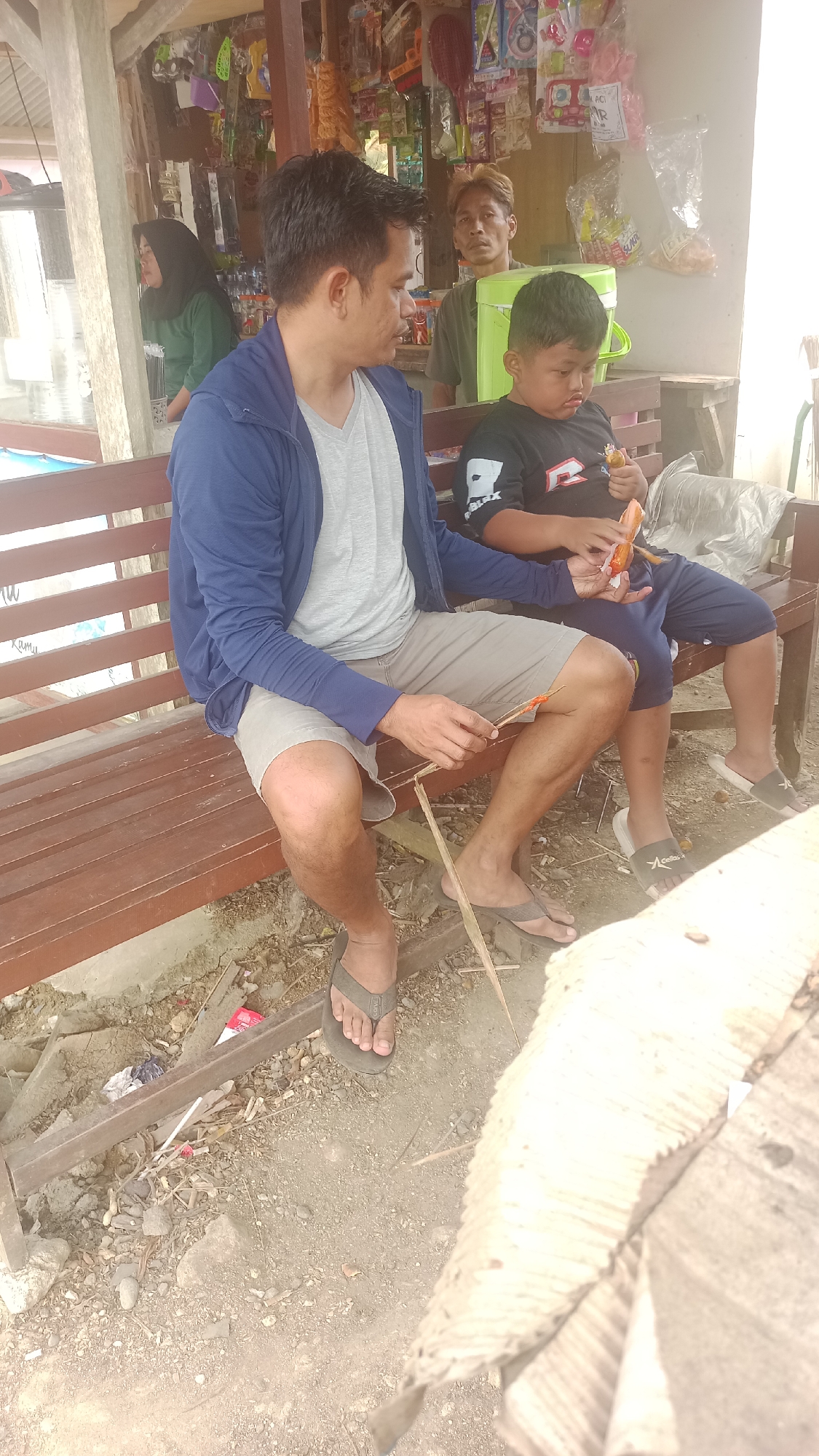 In Cibeber, Indonesia, a man and a child sit on a wooden bench in front of a small outdoor store. The man, wearing a blue jacket and gray t-shirt, is holding a stick with food on it. The child, in a black t-shirt, is also holding food. In the background, a woman in a green shirt and headscarf is visible inside the store, along with shelves stocked with various items. Another man stands behind a green cooler. The ground is covered in dirt and debris.