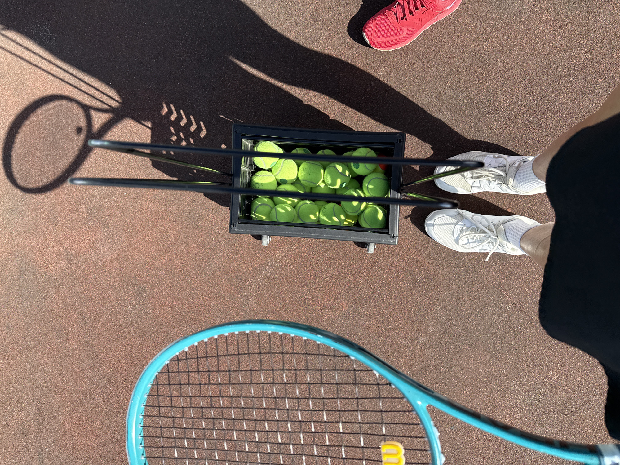 The image shows a top-down view of a tennis court in Chaikovsky, Russia. A turquoise tennis racket is visible on the left side of the frame. In the center, a black basket filled with approximately 20 bright green tennis balls sits on the court. Two pairs of feet are visible: one wearing white tennis shoes and socks, and another wearing red tennis shoes. The court surface is a reddish-brown color, characteristic of many outdoor tennis courts. Sunlight casts long shadows across the court, indicating a sunny day. The scene depicts a moment of preparation or pause during a tennis activity.