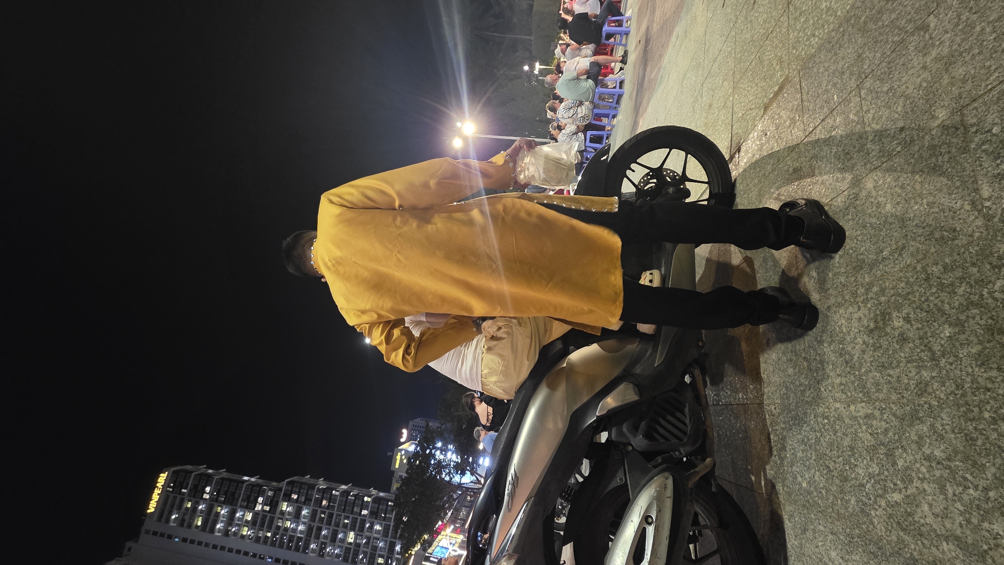 A man in a gold tunic and black trousers stands next to a parked silver and black motorbike. The man is holding a white plastic bag. The background shows buildings, city lights, and a group of people seated on chairs at tables. The image appears to be taken at night in Nha Trang Ward, Vietnam.
