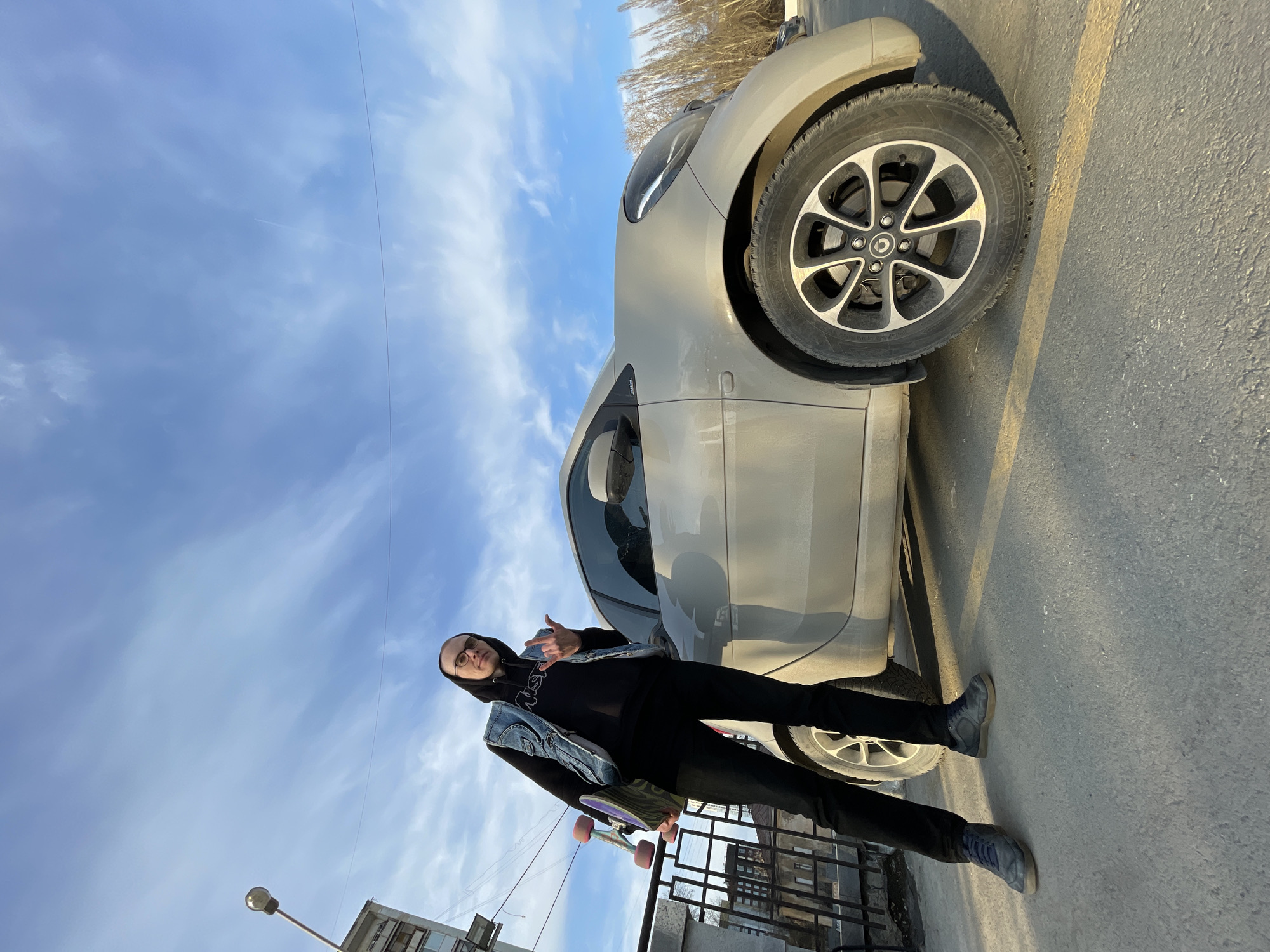The image depicts a man standing next to a light gray car on a street. The man is wearing a black hoodie, black pants, and a denim vest. He is also wearing sunglasses and holding a skateboard. He is making a gesture with his hand. The car is parked on the side of the road. In the background, there are buildings, including a block of flats, a fence, a lamp post, trees, and a cloudy blue sky. The photo appears to be taken in Yekaterinburg, Russia, based on visual evidence such as the car type, architectural styles, and available context. The surface is a paved street.