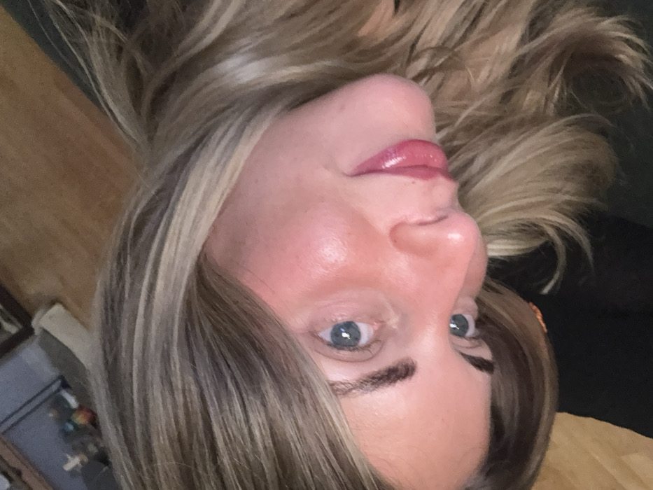 A woman's face fills the frame, tilted slightly to the side. She has fair skin, light brown hair with blonde highlights, and blue eyes. Her lips are full and glossed, and her eyebrows are dark and defined. The background is blurred and indistinct, with hints of furniture and fabric in dark colors. The lighting is soft and diffused, highlighting her features.