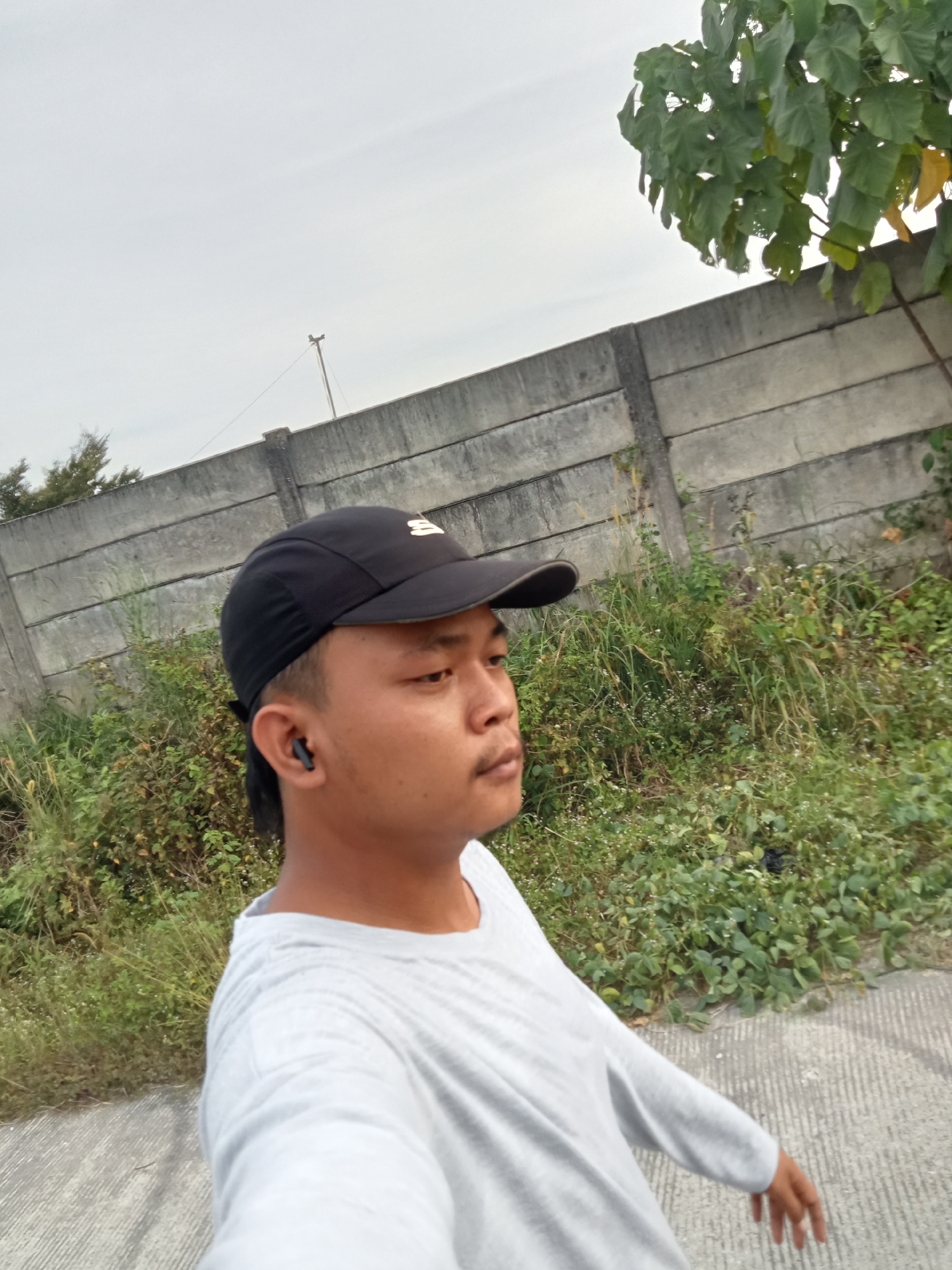 An Indonesian male wearing a gray long-sleeved shirt and a black baseball cap is captured in a side-profile, close-up shot. He is outdoors, walking on a paved path next to a concrete barrier that stretches horizontally across the frame. Lush green vegetation, including tall grasses and leafy plants, grows behind the barrier and to the right of the path. The sky above is overcast and pale. The man has short dark hair and is wearing a black earbud in his left ear. His gaze is directed forward and slightly to his right. The scene appears to be an urban or suburban environment in Teluk Kenidai, Indonesia.