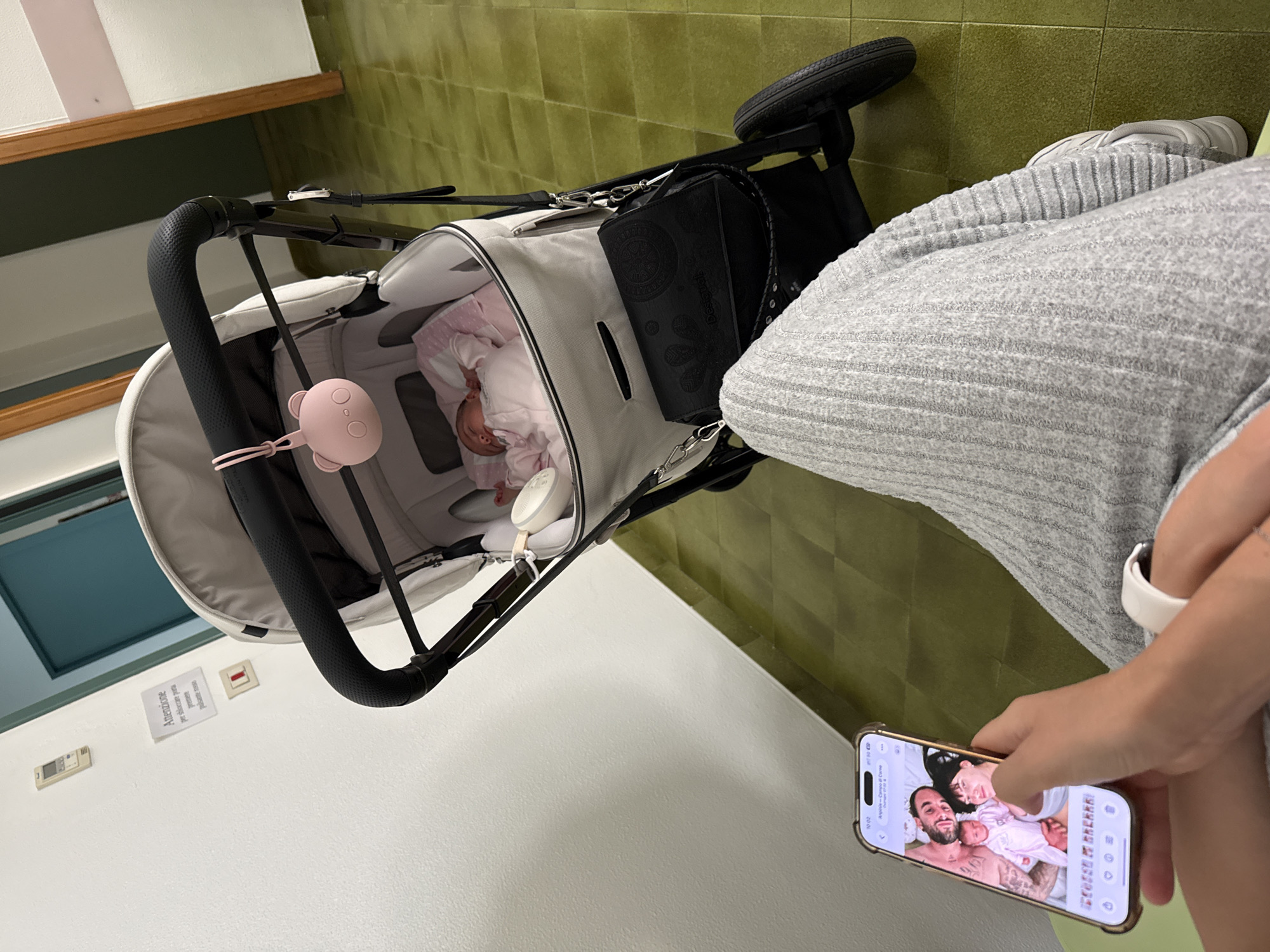 A woman is sitting in Aprilia, Italy, wearing gray knit pants and white sneakers, looking at a cell phone displaying photos of a baby, a man, and a woman. Next to her is a stroller with a baby inside. The room has green tiled floors and white walls with a sign written in Italian.
