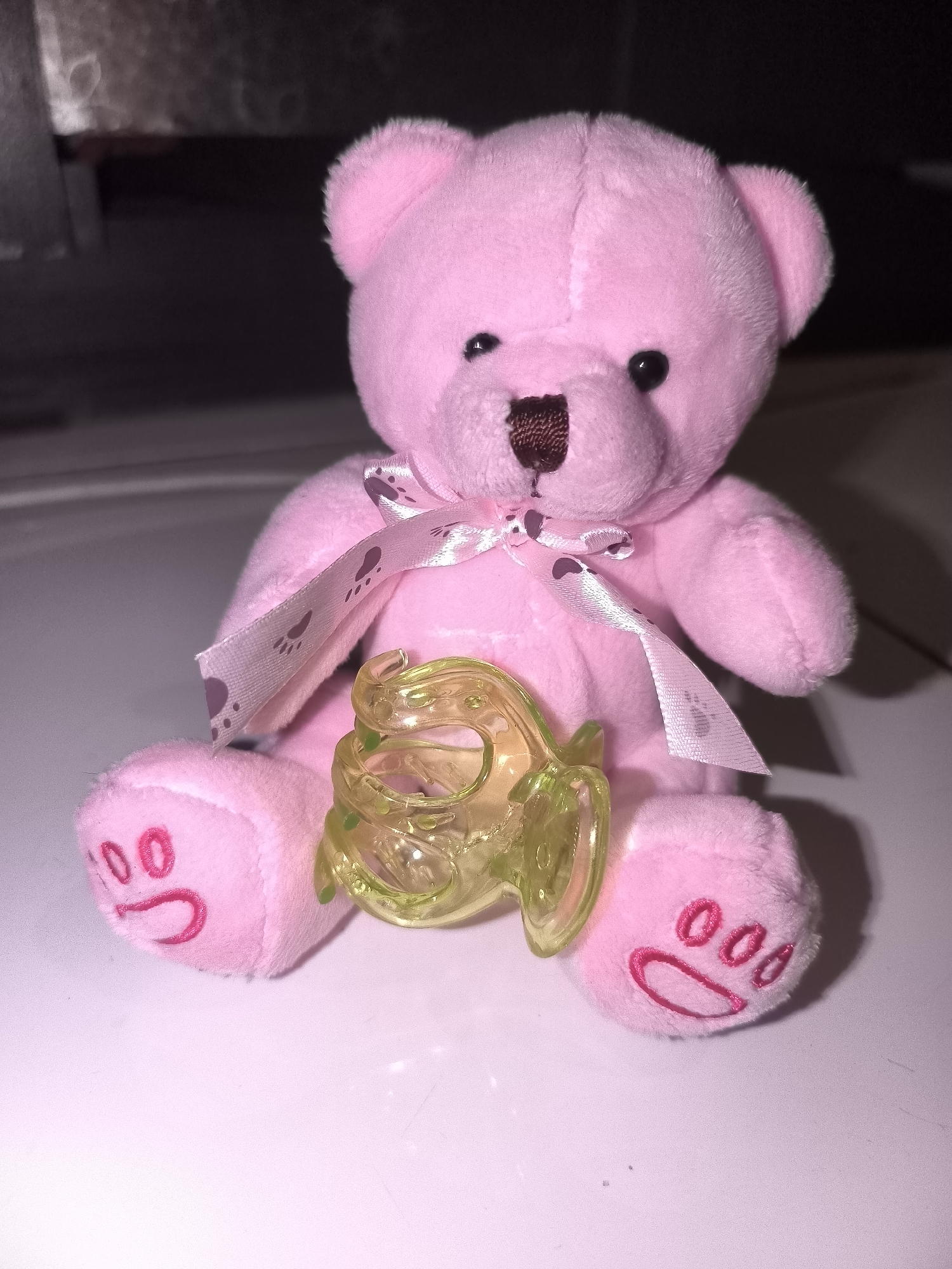 A pink teddy bear is sitting upright on a white surface, facing the viewer. The teddy bear has black eyes, a brown nose, and a pink ribbon tied around its neck with paw prints on it. A small, translucent yellow object with green dots is placed in front of the teddy bear. The teddy bear's paws have pink paw print designs. The background is dark and out of focus. The image was taken on a white surface, potentially a table or floor, indoors. No specific location in North Jakarta, Indonesia, can be determined from the image.