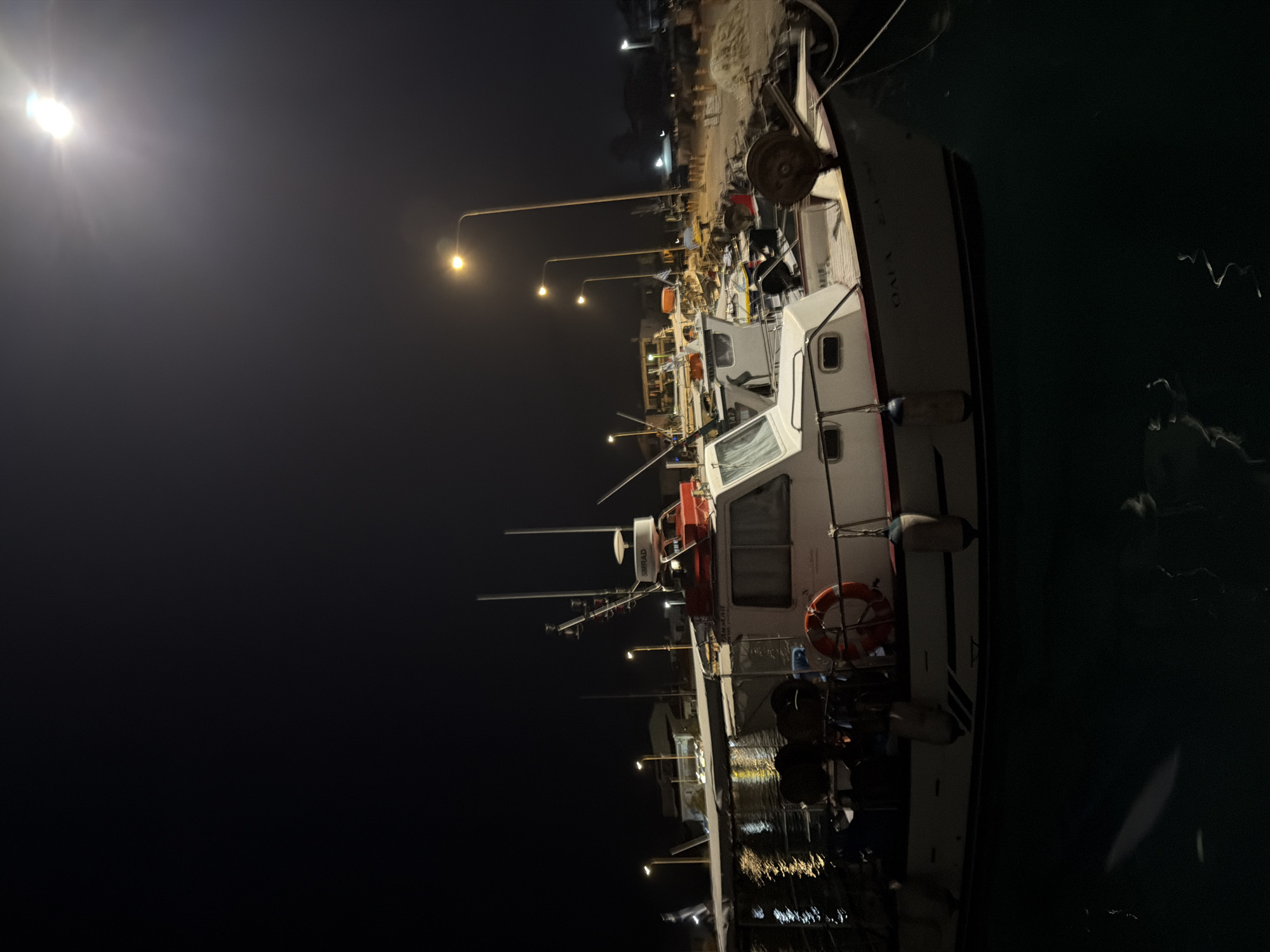 A night shot captures a moored fishing boat in the Municipal Unit of Alyzia, Greece. The boat, predominantly white with a red trim, features a cabin structure and is equipped with various fishing gear, including nets and ropes. A life preserver ring is visible on its side. The boat is docked in dark water, with reflections from nearby lights shimmering on the surface. Several streetlights illuminate the scene, casting a warm glow and creating long shadows. In the background, other boats are moored, and structures of buildings are faintly visible. The sky is dark, with a bright, possibly moonlit, sphere dominating the upper right corner of the image. The overall atmosphere is that of a quiet, working harbor at night.