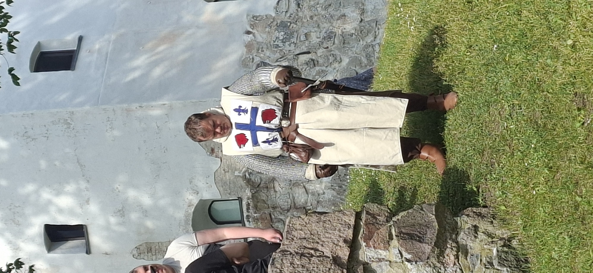 A person dressed as a medieval knight stands on a grassy area in front of a stone wall and a white building. The knight wears a chainmail shirt, a white surcoat with a blue cross and red embellishments, brown leather boots, and pouches attached to a belt. The person is looking down. In the background, there is a white building with small windows. The location is the grounds of Turku Castle in Turku, Finland.