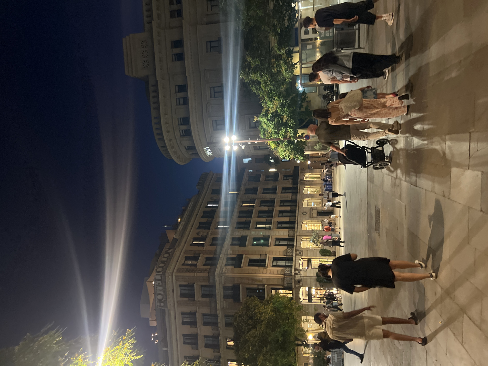 A nighttime scene depicts people walking in a plaza in Barcelona, Spain, with buildings on either side. In the foreground, two men and a woman walk away from the camera. The man on the left wears a striped short-sleeved shirt and light-colored shorts. The woman in the middle wears a patterned jumpsuit and carries a bag over her shoulder. The man to the right of the woman wears a dark green shirt and tan pants, and is pushing a stroller. Further back, a group of people are seen walking, some with shopping bags, and others talking amongst themselves. The buildings flanking the plaza are illuminated, with one on the left featuring the sign "HOTEL COLON" and another on the right with the "BBVA" logo. Trees are visible between the buildings and the plaza, and streetlights cast beams of light across the scene. The ground is paved with stone tiles.