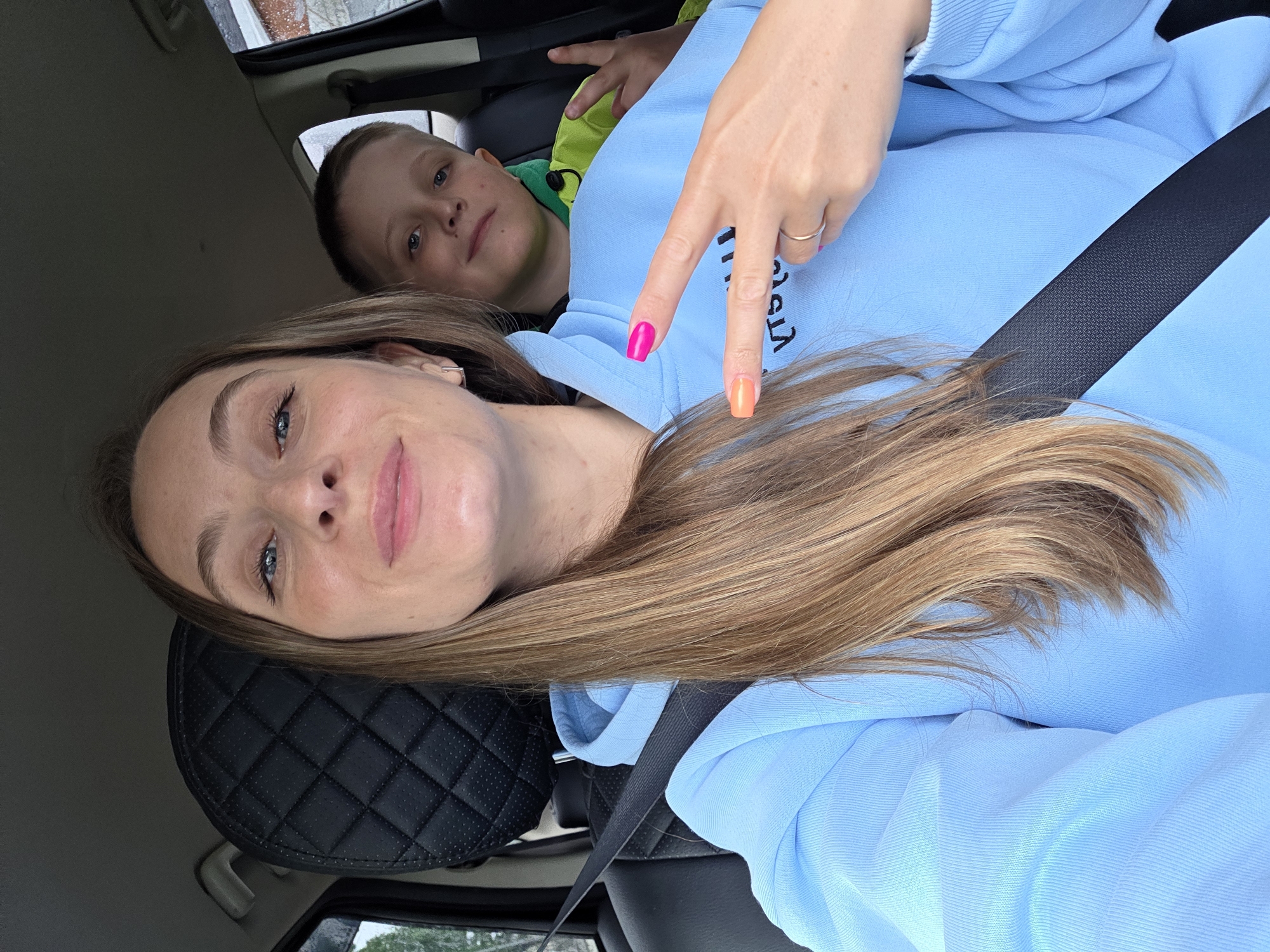 A selfie taken inside a vehicle shows a woman in the foreground and a child behind her.
The woman, with long, light brown hair and blue eyes, wears a light blue hooded sweatshirt and a black seatbelt across her chest. Her left hand is raised, forming a peace sign, with fingernails painted hot pink on the index and middle fingers, orange on the ring finger, and light pink on the pinky. A thin silver ring is on her left ring finger, and a small rectangular silver earring is visible in her left ear. She has a slight smile and looks directly at the camera.
Behind her, a young boy with short, light brown hair and blue eyes wears a bright lime green jacket. He also displays a peace sign with his right hand, partially obscured. He smiles faintly and looks towards the camera.
The vehicle interior features dark seating, with a quilted black headrest visible behind the woman. The outside environment, seen through the car windows, appears to be an overcast day with blurred trees and general outdoor scenery visible.