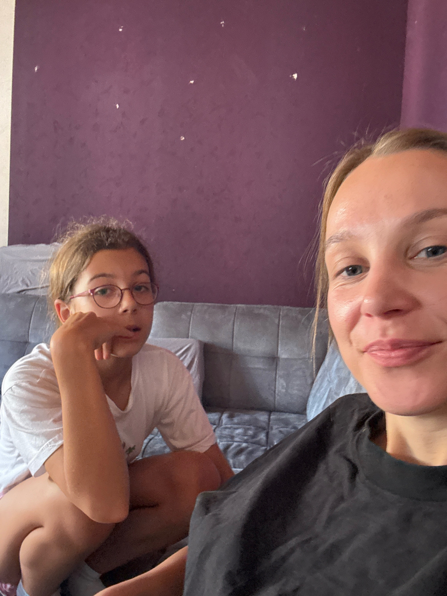 This is a selfie-style, medium shot photograph of two individuals, likely a mother and daughter, seated indoors against a purple textured wall. On the left, a young girl with blonde hair and glasses is seen from the chest up. She is wearing a white t-shirt and her left arm is bent, with her hand resting near her chin in a thoughtful or pensive pose. Her eyes are looking slightly to her left. On the right, a woman, appearing to be an adult with blonde hair, is captured from the shoulders up, close to the camera. She is wearing a black t-shirt and is smiling slightly, looking towards the camera. The background consists of a grey tufted sofa. The overall lighting suggests an indoor setting, possibly a living room. This image was taken in Überherrn, Germany.