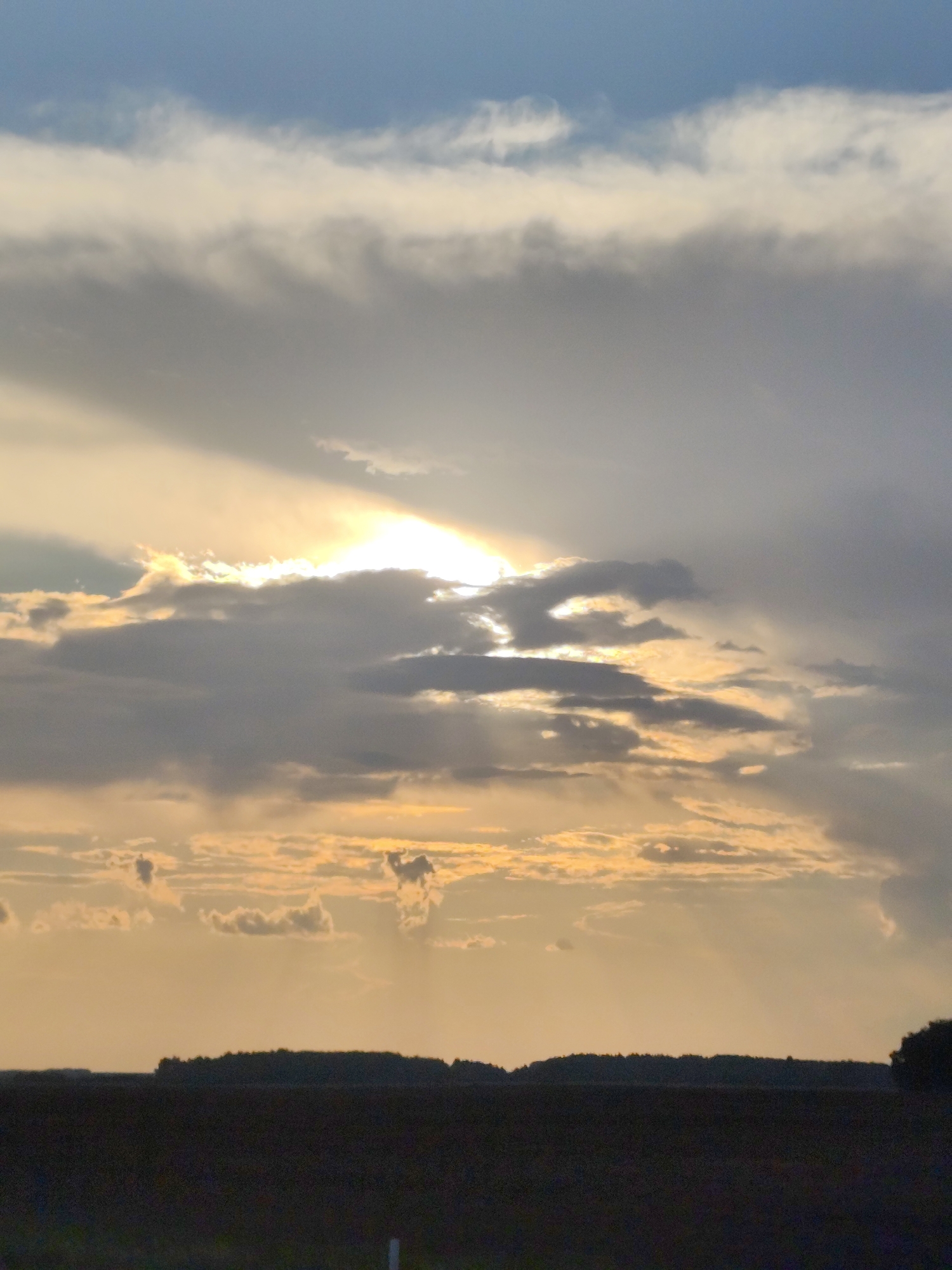 The image shows a wide-angle view of a sky with dramatic cloud formations and a muted sunset. The sun is partially obscured by clouds, creating rays of light that pierce through the atmospheric layers. The lower portion of the image depicts a dark, silhouetted landscape, likely a field or treeline, against the lighter sky. The overall mood is serene and somewhat dramatic due to the lighting and cloud cover. There are no people, objects, or actions visible in the image. This scene is a natural landscape, not specific to any urban location.