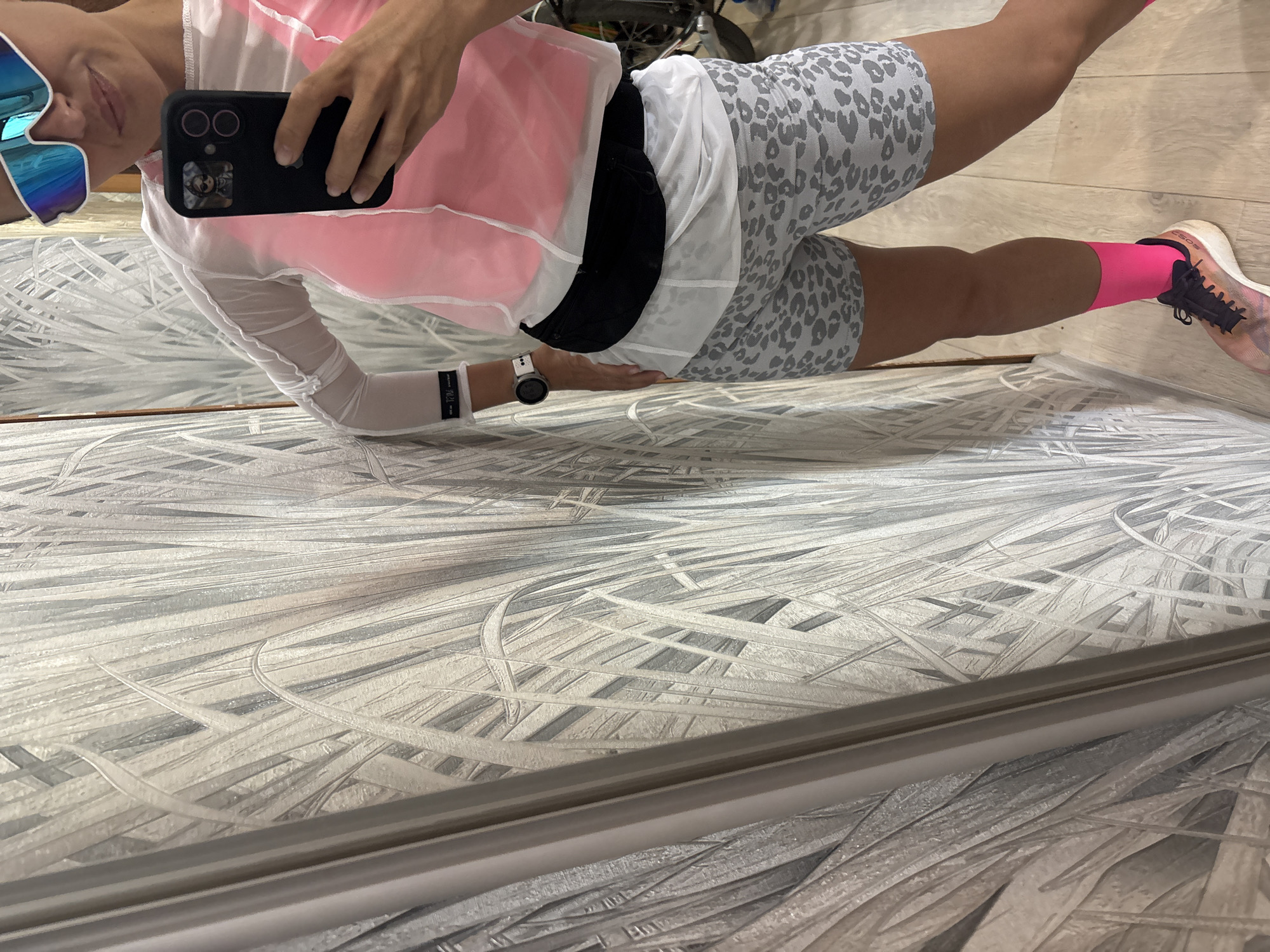 A person is taking a selfie in front of a mirror. The person is wearing colorful sunglasses, a white top with a pink sleeveless shirt, leopard print shorts, and pink socks with sneakers. The person is holding a black smartphone. The background appears to be a patterned wall or flooring. A blurred object, possibly a walker or chair, is visible behind the person. The location is not identifiable as Chaikovsky, Russia from the visual information provided.