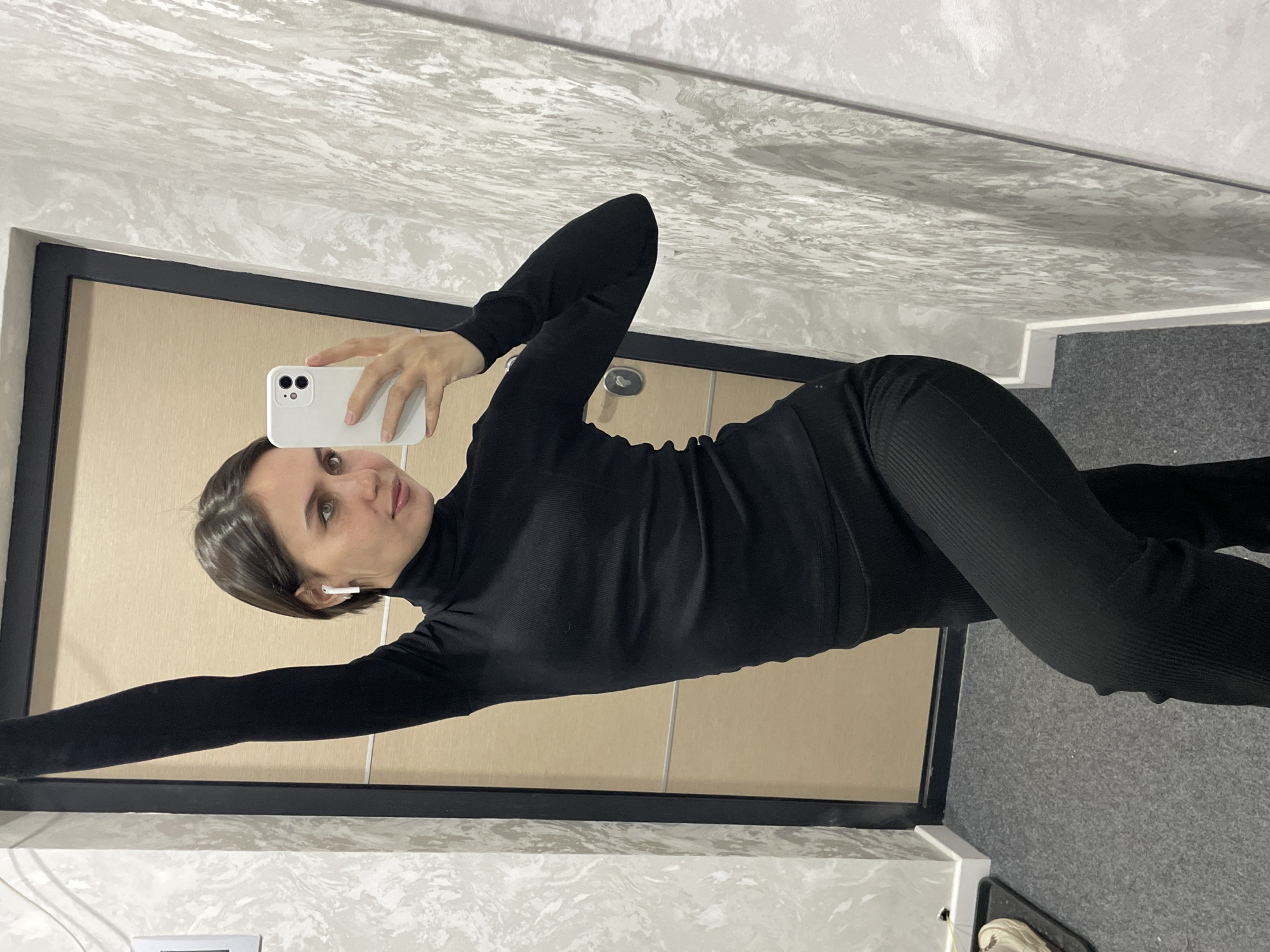 This image captures a female individual taking a mirror selfie indoors in the city of Izhevsk, Russia. The subject, a young adult with short dark hair, is dressed in a black ribbed long-sleeve turtleneck top and matching black ribbed pants. She has a white wireless earbud inserted in her right ear. In her left hand, she holds a white smartphone with dual rear cameras, which is directed towards her reflection in the mirror to capture the image. Her right arm is extended vertically upwards. The subject's body is angled with her left knee slightly bent, and she is looking directly into the camera's reflection.
The background elements include a light-colored wooden door with black trim and a silver handle, visible as a reflection behind the subject. The walls flanking the mirror exhibit a grey and white marbled texture. The floor is covered with a dark grey carpet or mat. Partially visible on the far left of the floor, atop the mat, is a light-colored shoe or slipper. A white electrical outlet or switch is mounted on the wall to the left of the mirror.
