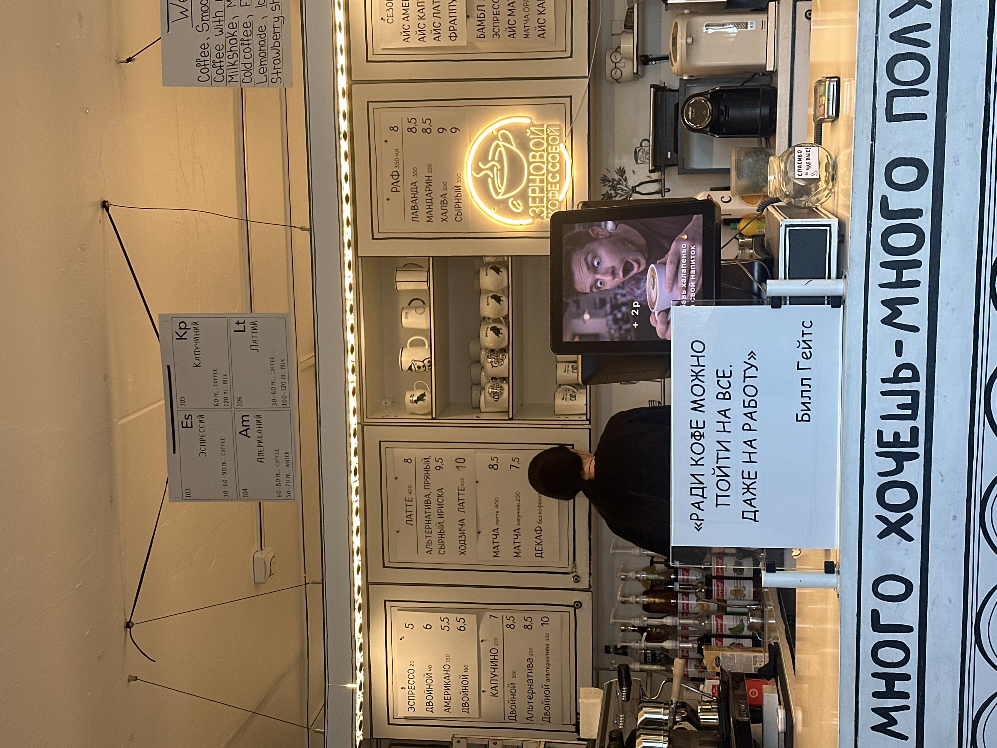 The image shows a coffee shop counter area. In the foreground, a sign with large, bold, black Russian text reads "МНОГО ХОЧЕШЬ - МНОГО ПОЛУ" which translates to "You want a lot - you get a lot." Behind this sign, on the counter, is a transparent glass jar with a handwritten note on it that says "СПАСИБО ЧАЕВЫЕ!" meaning "Thank you for the tips!". Next to the jar is a credit card reader. A digital screen displays a man's face with a surprised expression holding a cup of coffee and the text "+ 2p" and "свой напиток" (your drink).
Above the counter, menus are displayed on the wall. The leftmost menu lists coffee drinks like "ЭСПРЕССО" (Espresso), "ДВОЙНОЙ" (Double), "АМЕРИКАНО" (Americano), and "КАПУЧИНО" (Cappuccino) with their corresponding prices and sizes. The middle menu lists "ЛАТТЕ" (Latte), "АЛЬТЕРНАТИВНАЯ, ПРЯНЫЙ, СЫРНЫЙ, ИРИСКА" (Alternative, Spicy, Cheesy, Toffee), "ХОДЗИЧА ЛАТТЕ" (Khodzhcha Latte), "МАТЧА" (Matcha), and "ДЕКАФ" (Decaf). The rightmost menu has headings in Russian, but the text is largely illegible.
A neon sign in the shape of a steaming coffee cup with the text "ЗЕРНОВОЙ КОФЕ С СОБОЙ" (Takeaway Coffee Beans) is illuminated.
In the background, a person with dark hair is partially visible, appearing to be an employee behind the counter. Various coffee-making equipment and bottles of syrups are visible on shelves and the counter. The environment appears to be a modern, well-lit coffee shop. The presence of Russian text on menus and signs suggests a location where Russian is spoken, such as Minsk, Belarus.