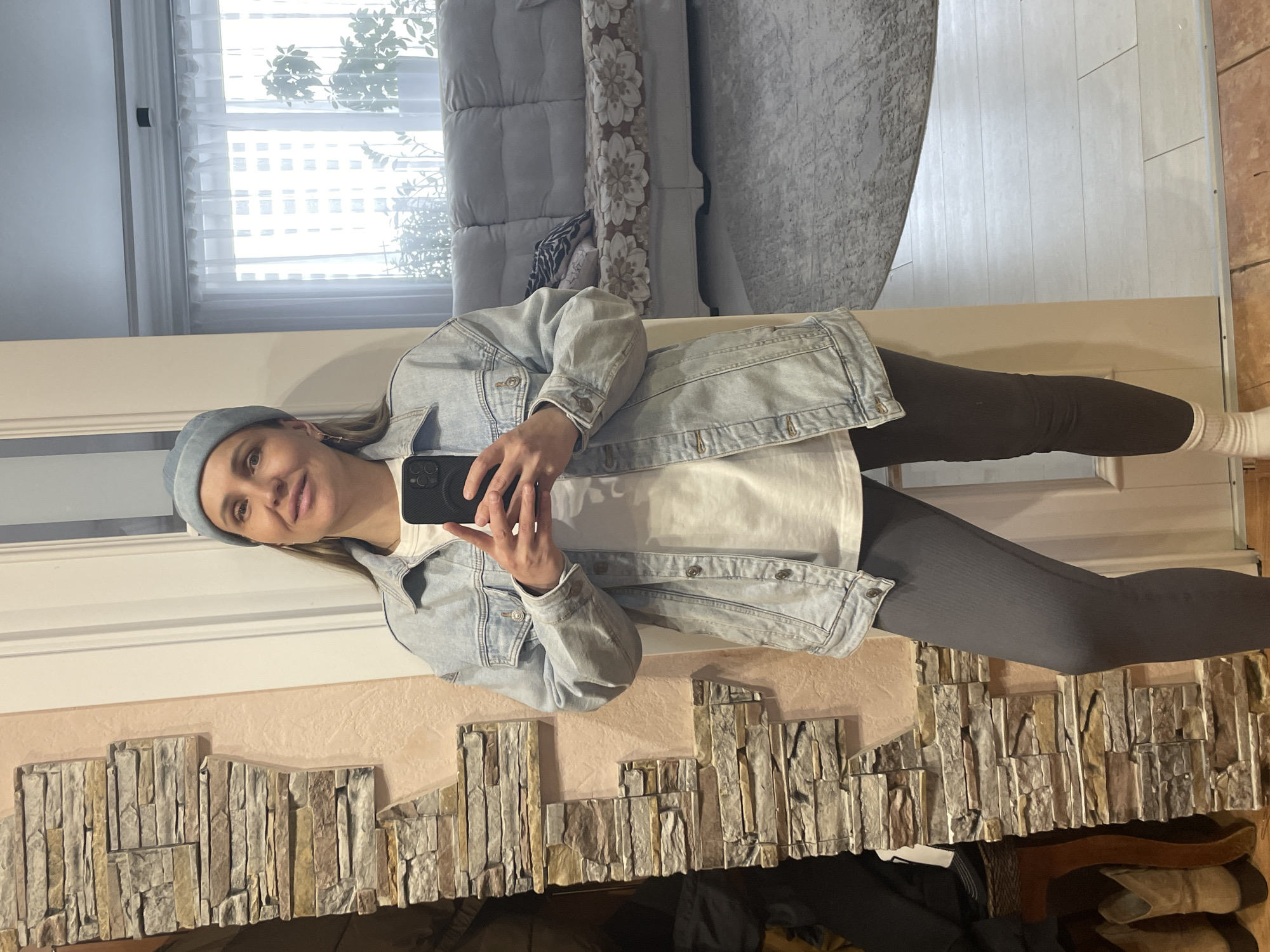 A young woman wearing a light blue hat and denim jacket, a white t-shirt, and gray leggings is taking a selfie in a room with a mirror. The mirror reflects a couch, a window with curtains, and a wall with a decorative stone pattern. Various clothing items and shoes are visible beneath the mirror. The location is in Chaikovsky, Russia.