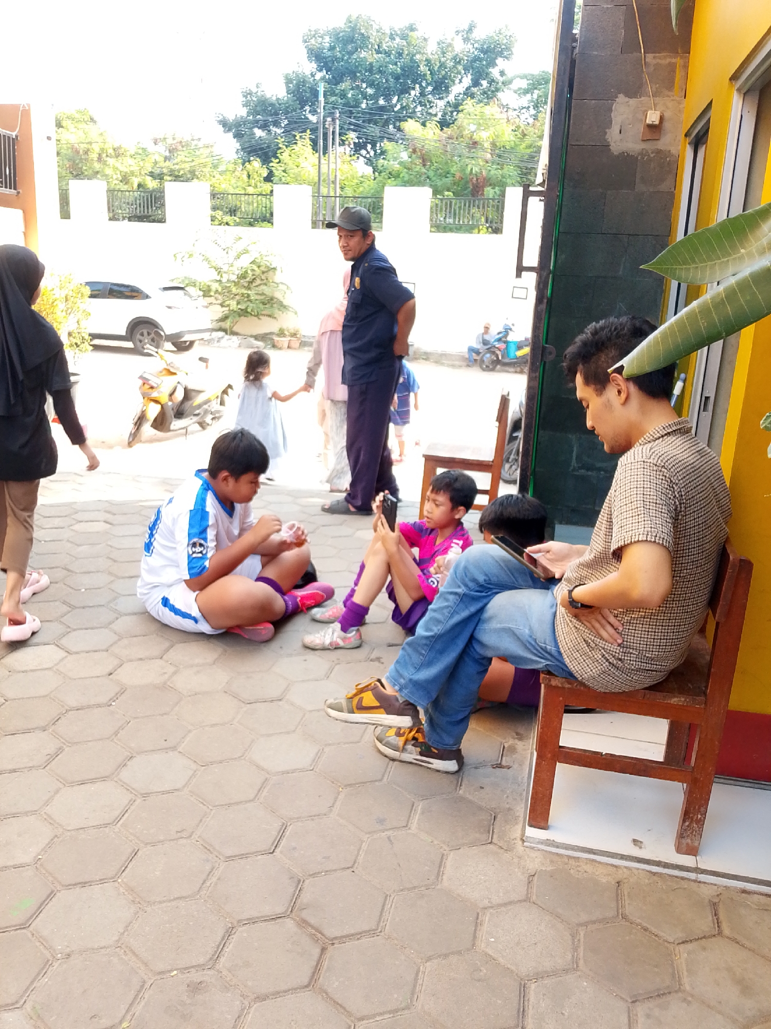 In the city of Central Jakarta, Indonesia, a group of people are gathered outdoors on a paved, hexagonal-patterned surface. A man wearing a plaid shirt and jeans sits on a wooden chair, looking at his phone. To his left, a boy in a white and blue soccer jersey is sitting on the ground, also engaged with a small object. Further left, another boy in a pink shirt is seated on the ground, looking at his phone. In the background, a man in a dark blue uniform and a cap stands facing away from the viewer. Several other individuals, including a woman in a hijab and a child in a light blue dress, are also present, with a yellow scooter visible behind them. Trees and a white fence are in the distance. The overall scene appears to be a casual gathering outdoors.