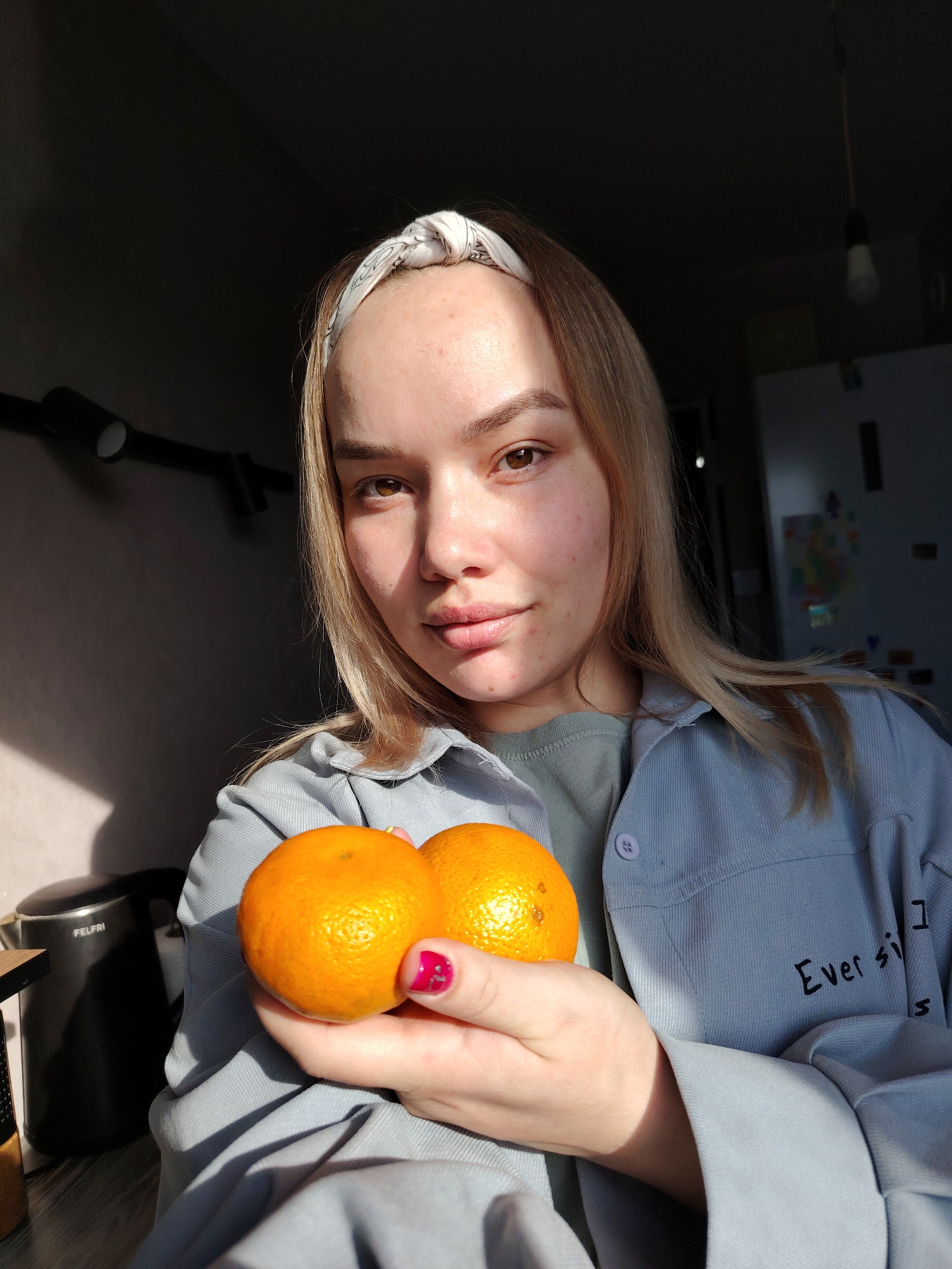A fair-skinned woman is holding two mandarin oranges in her left hand. She is wearing a patterned headband, a light blue corduroy shirt over a green shirt, and her fingernails are painted pink. The location is Марковский, Russia. There is a kettle visible to the left, and a refrigerator can be seen in the background. Sunlight illuminates the left side of her face.