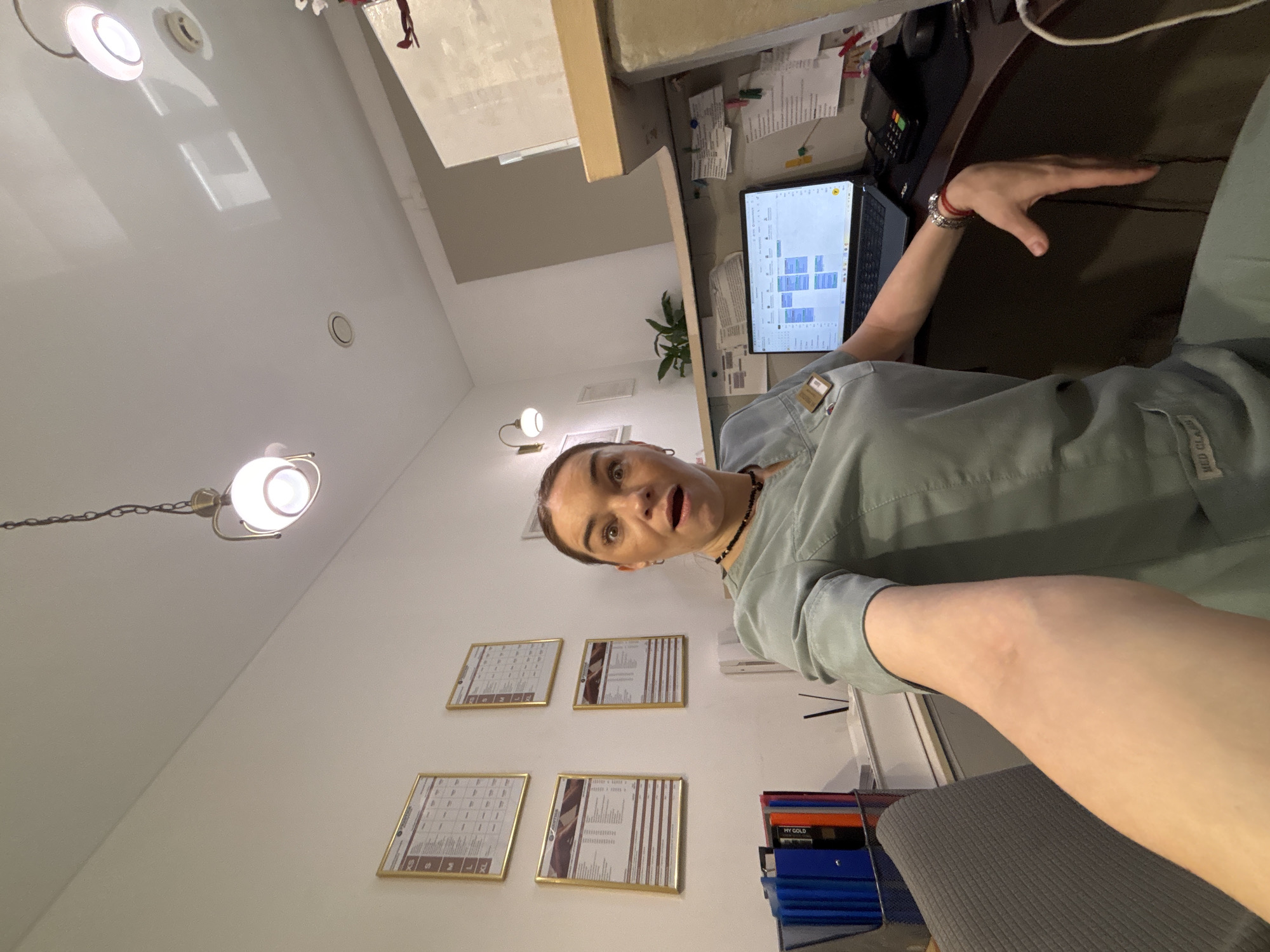 A woman is in an office setting. She is standing behind a desk, on which there is a laptop, a phone, and several documents. There are framed calendars on the wall behind her. The room is lit by overhead fluorescent lights and a decorative hanging lamp. The location is in the city of Unknown, Unknown.