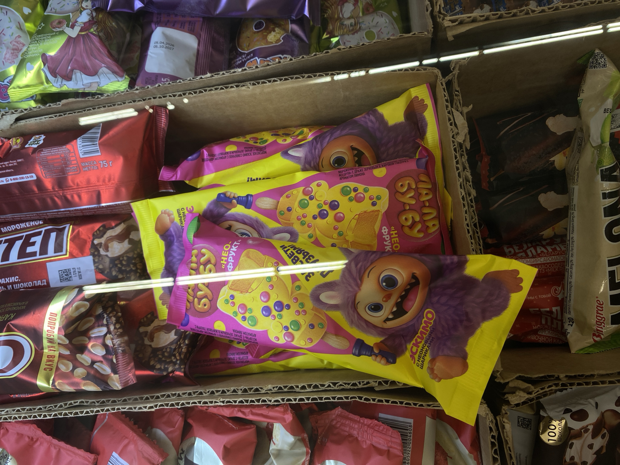 The image depicts a close-up, top-down view of a cardboard box filled with various ice cream and confectionery products. The products are predominantly packaged in brightly colored wrappers, with a focus on yellow, pink, and red. Several of the items are ice cream bars with illustrated characters. One prominent item is a yellow wrapper featuring a cartoon monster character holding an ice cream. Other wrappers display images of ice cream cones and fruit. In the background, there are additional stacks of packaged goods. The lighting suggests an indoor environment, likely a retail setting such as a supermarket or convenience store, with vertical fluorescent lights visible along the right edge of the frame, indicating a refrigerated display case. The overall scene is one of retail product display, with an emphasis on visually appealing packaging, especially for children. There are no people or animals visible in the image. The setting is identified as Kuzmolovskiy, Russia.