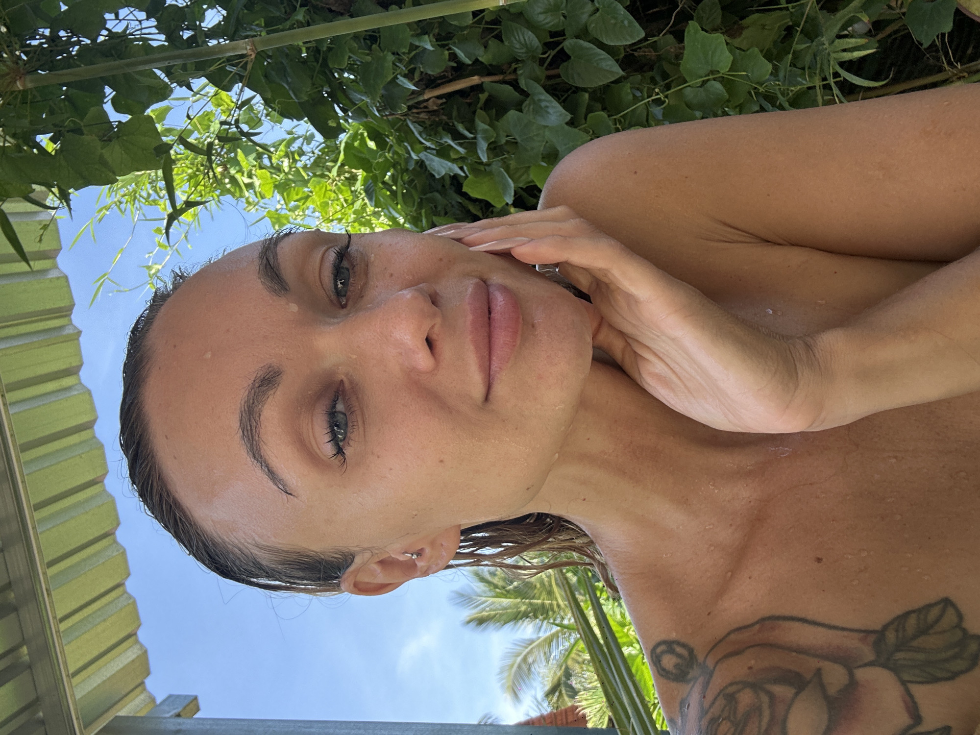 A woman with fair skin and wet hair looks at the camera while her left hand is touching her face. She has a tattoo of a rose on her left shoulder. Behind her are leafy green plants. In the distance are palm trees. To her left, there is a green corrugated metal roof. The image was likely taken outside, in the city of Mũi Né, Vietnam.