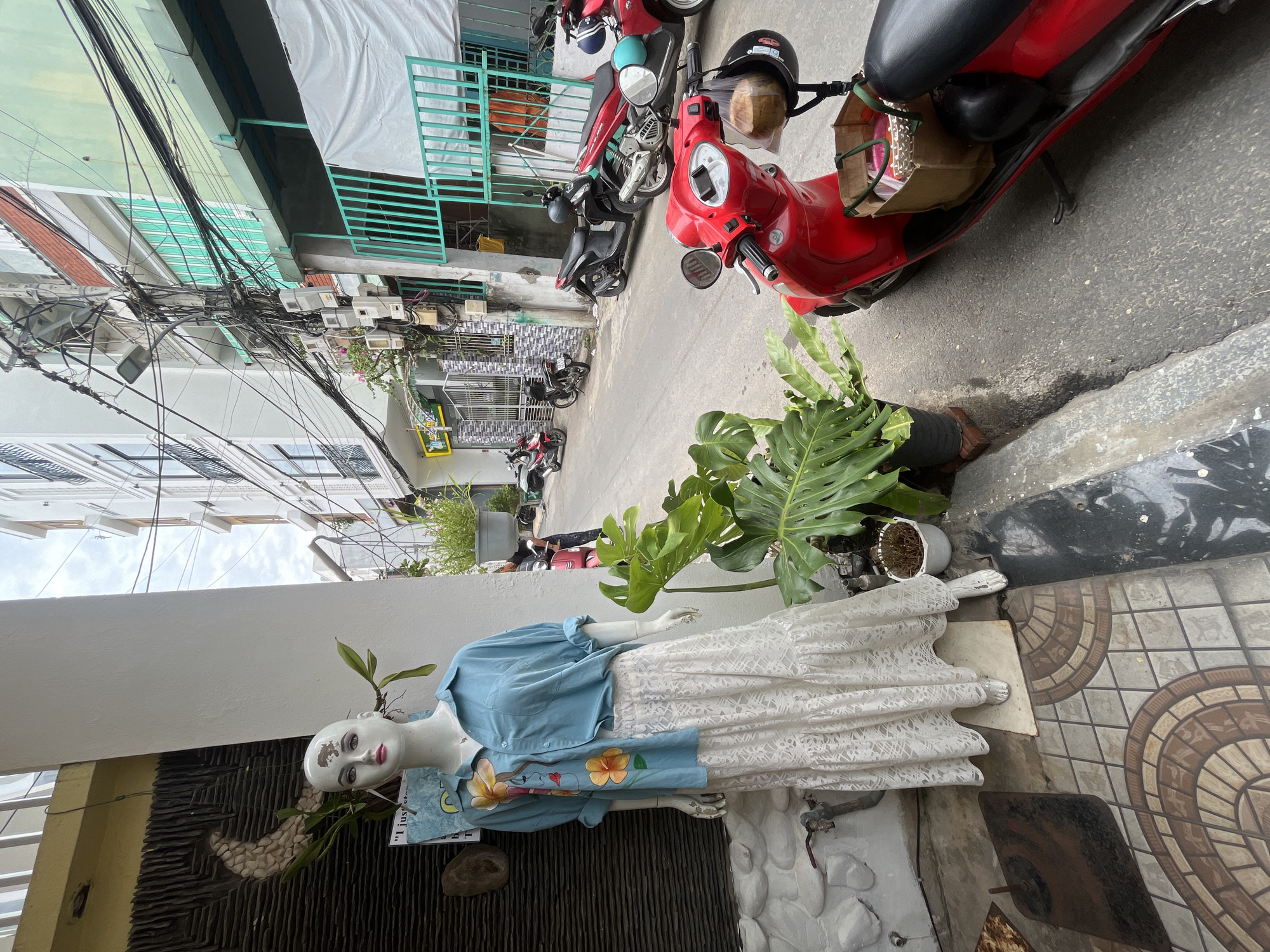 The image depicts a street scene in the city of Đà Nẵng, Vietnam, likely in a commercial area. In the foreground, a white mannequin is displayed outside a shop, wearing a light blue, oversized button-down shirt with a floral design and a long, white lace skirt. The mannequin stands on a tiled floor with decorative patterns. To the right of the mannequin, a red scooter is parked, with a coconut and a black helmet hanging from its handlebars, and a canvas bag with items inside placed on the seat. Beyond the mannequin and scooter, the street continues, lined with buildings of varying heights and architectural styles. Many buildings have balconies and are adorned with numerous electrical wires and conduits, indicative of a densely populated urban environment. Several other motorbikes and scooters are parked along the street and in the background. Lush green plants in black pots are placed on the sidewalk, adding a touch of nature to the urban setting. The sky is overcast, suggesting a cloudy day.