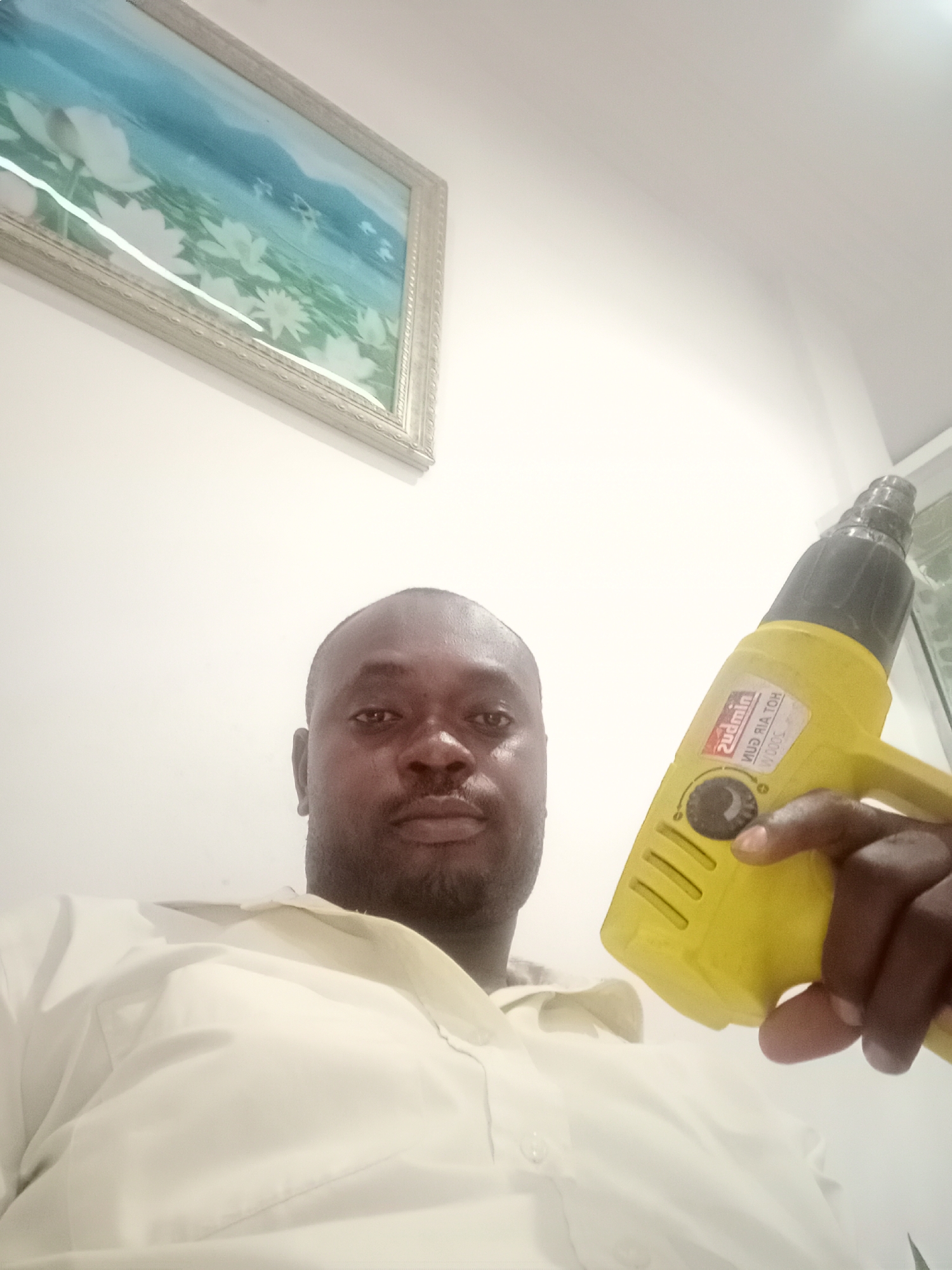 A man is holding a yellow "Zudmin Hot Air Gun" tool in his right hand. The man is wearing a white shirt. In the background, there is a framed picture hanging on a white wall. The location is not specified as Gwarinpa, Nigeria in the image, so no mention of city and country are included in this description.