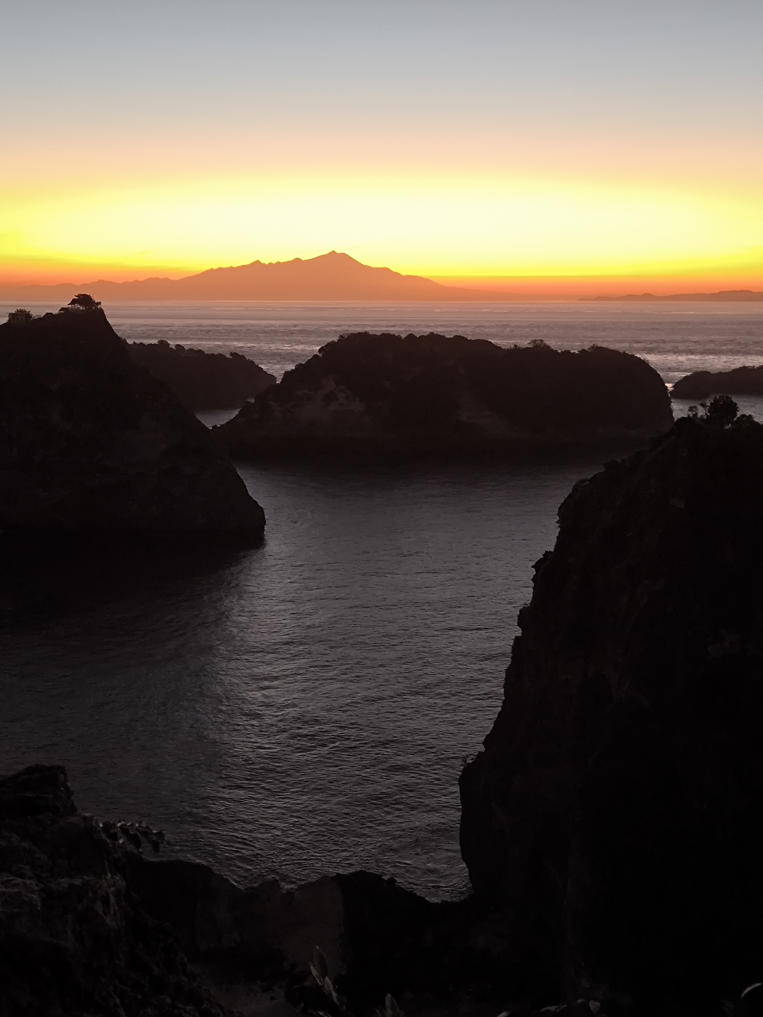 The image depicts a coastal view in Nusa Penida, Indonesia, captured during sunrise or sunset. The foreground consists of dark, rocky cliffs framing a body of water. Several small, rocky islands dot the water, with one featuring a cluster of trees on top. In the distance, the silhouette of a mountain range is visible against a vibrant sky gradient of orange, yellow, and pale blue. The water is dark with ripples, and the overall scene is tranquil.