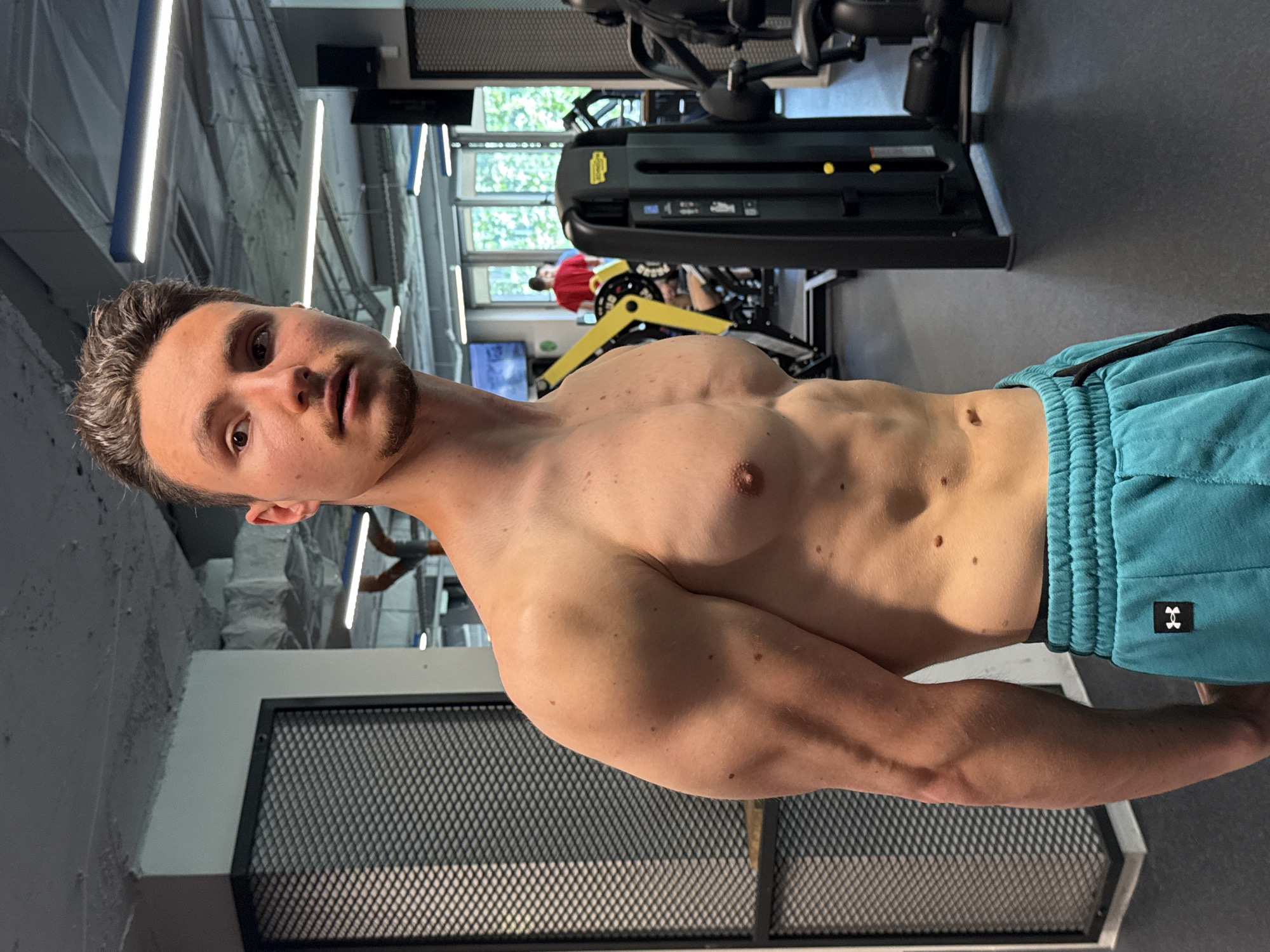 A shirtless man is the primary subject of the image. He is wearing teal athletic shorts and is standing in what appears to be a gym. The man has a medium build, short brown hair, and light skin. Gym equipment can be seen in the background. Based on the details visible in the background, the location of this image is in a gym located in Sofia, Bulgaria. The specifics cannot be deduced further.