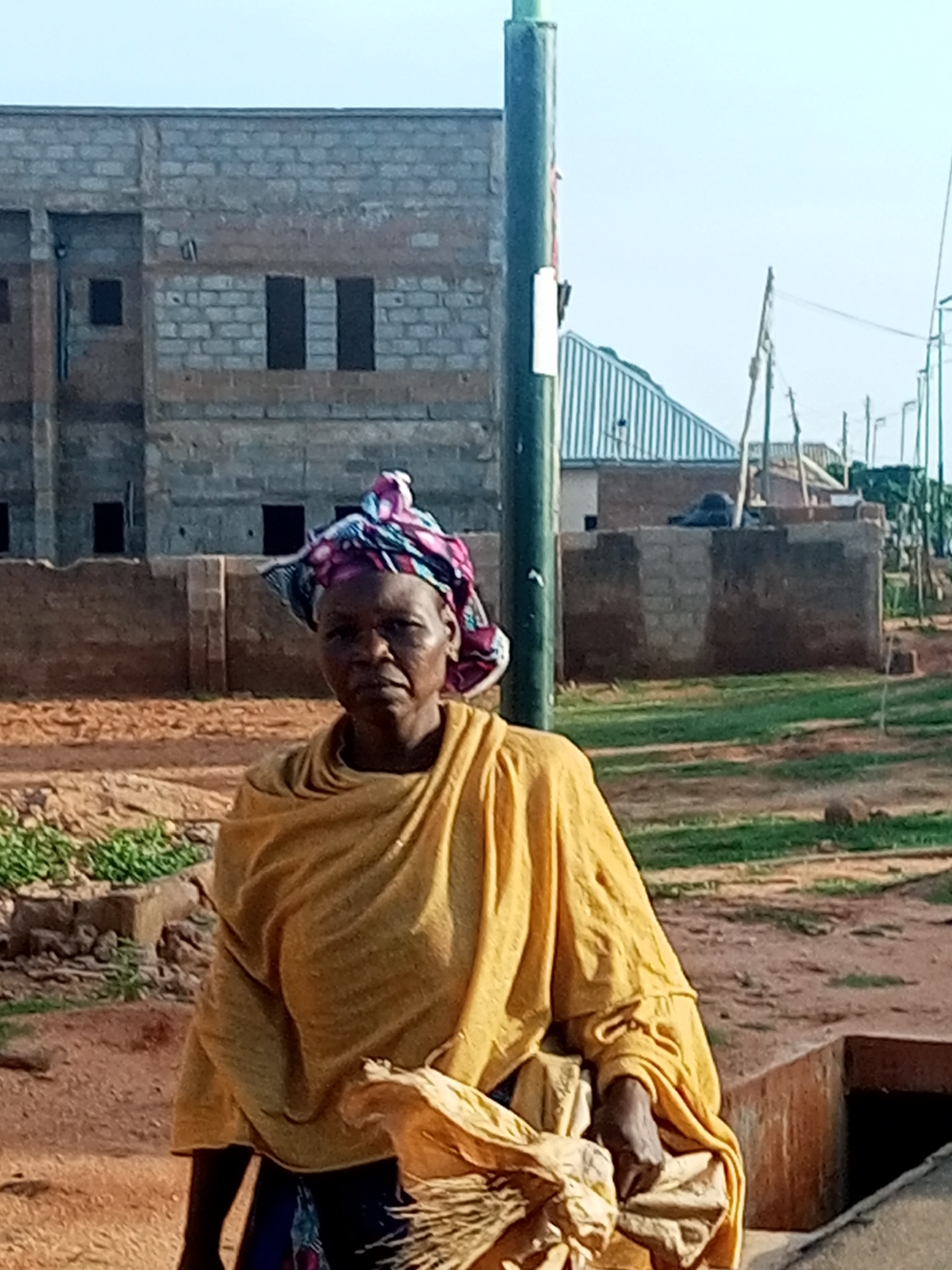 A woman stands in an outdoor urban environment. She is looking directly at the camera with a neutral expression. She is wearing a yellow garment and a multi-colored headscarf. She is holding a bundle of what appears to be straw in her right hand. Behind her, there is a partially constructed building made of concrete blocks, a utility pole, and a dirt ground surface with some vegetation. The scene is lit by natural daylight. The location is Tempure, Nigeria.