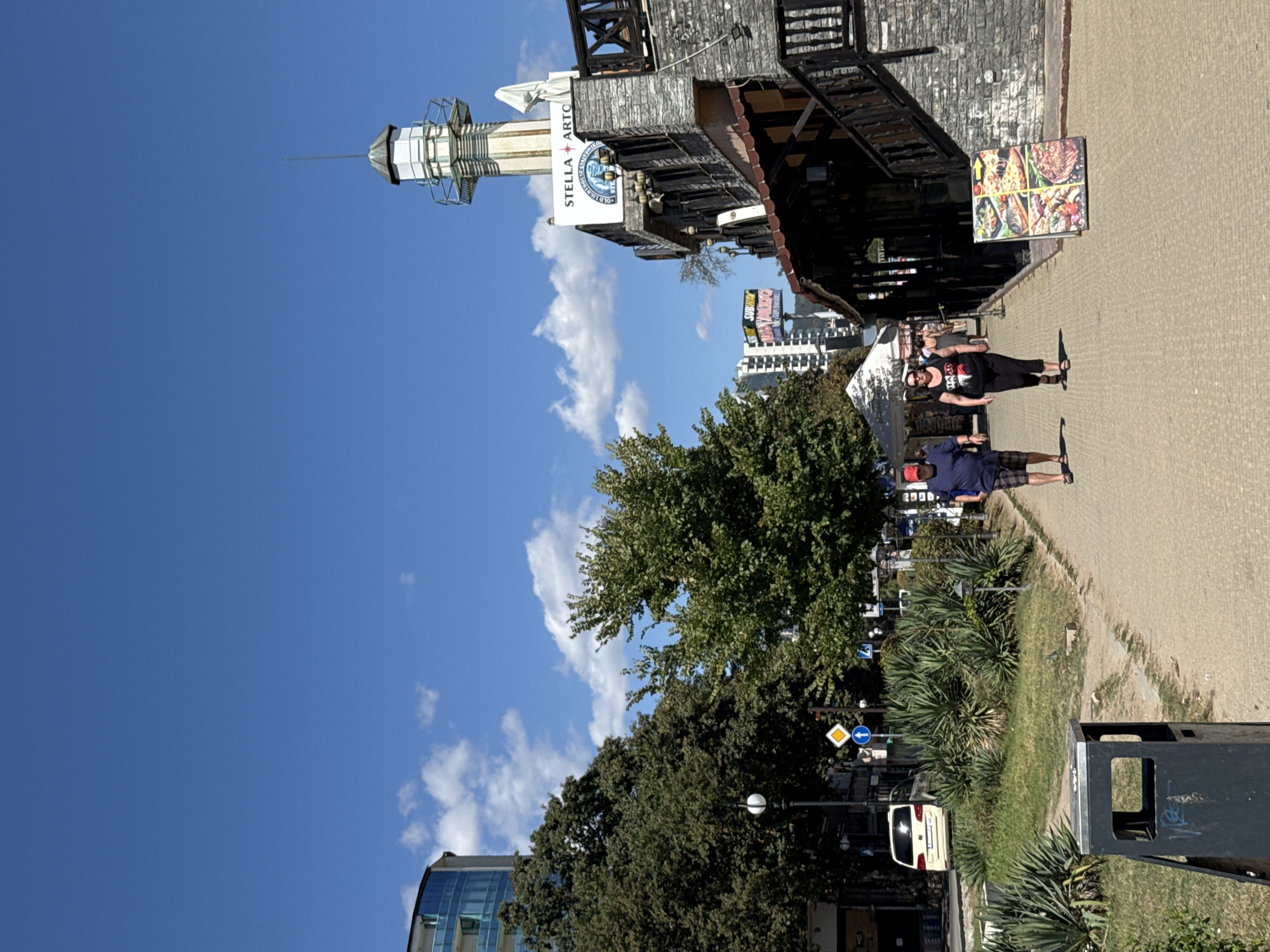The image captures a sunny day along a paved walkway in Varna, Bulgaria. On the right, a two-story building with a stone facade is visible, topped by a lighthouse-like structure. A sign reads "STELLA ARTO." In front of the building, a display board showcases pictures of food, including pizza and meat dishes. To the left of the walkway, lush green trees and shrubs line the path, with a modern glass-fronted building in the background. A few people are seen walking on the path. One man in a blue shirt and plaid shorts, wearing a red cap, walks towards the foreground. A woman in a black tank top and black leggings walks beside him. Further down the path, more people and tents are visible. A white van is parked on the street to the left, behind a grassy area with yucca plants. Traffic signs and streetlights are also present along the roadside. The sky is clear blue with scattered white clouds. The overall scene suggests a vibrant, potentially tourist-oriented area in Varna, likely near the coast given the presence of the lighthouse structure.