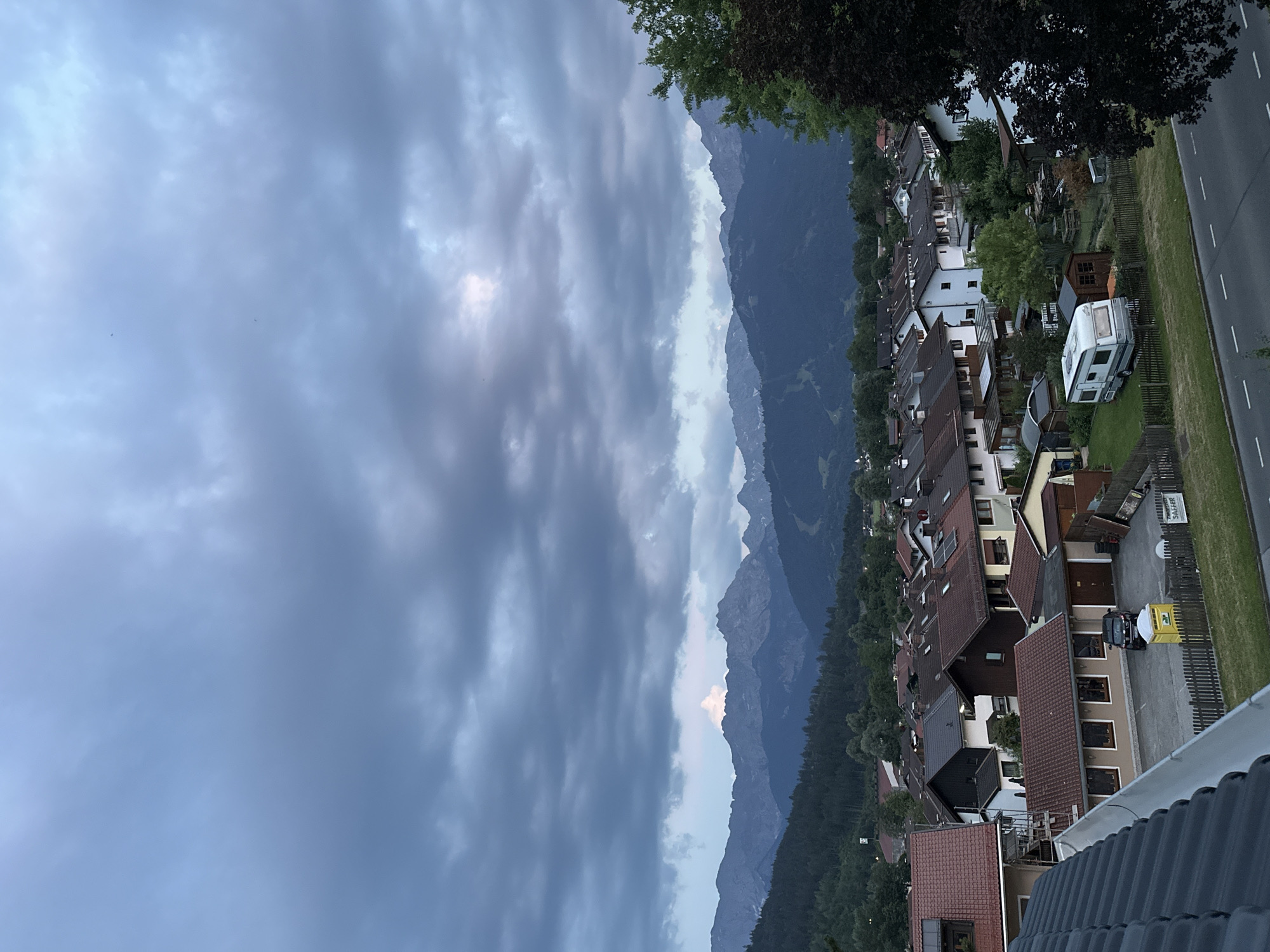 The image depicts a twilight scene in Garmisch-Partenkirchen, Germany, looking out over a residential area towards a mountain range. The sky is filled with heavy, dark clouds, with a hint of lighter sky peeking through on the horizon. Below the sky, a dense collection of houses with red and dark-colored roofs is visible. In the foreground, part of a dark-tiled roof is prominent. A road with white lane markings runs along the bottom right of the image.
Among the houses, several details are discernible: a white caravan is parked on a grassy area with a wooden fence. A small wooden shed is situated next to the caravan. A yellow portable toilet is visible in front of a fence in the lower middle section of the image, with a dark-colored car parked nearby. Further back, more houses and trees create a layered effect, leading to the backdrop of the dark, forested foothills of the mountains. The mountains themselves are a deep blue-gray, with jagged peaks. A small, white, cloud-like formation is visible above one of the mountain peaks. The overall atmosphere is one of approaching evening or a storm, with muted light and a sense of quiet.