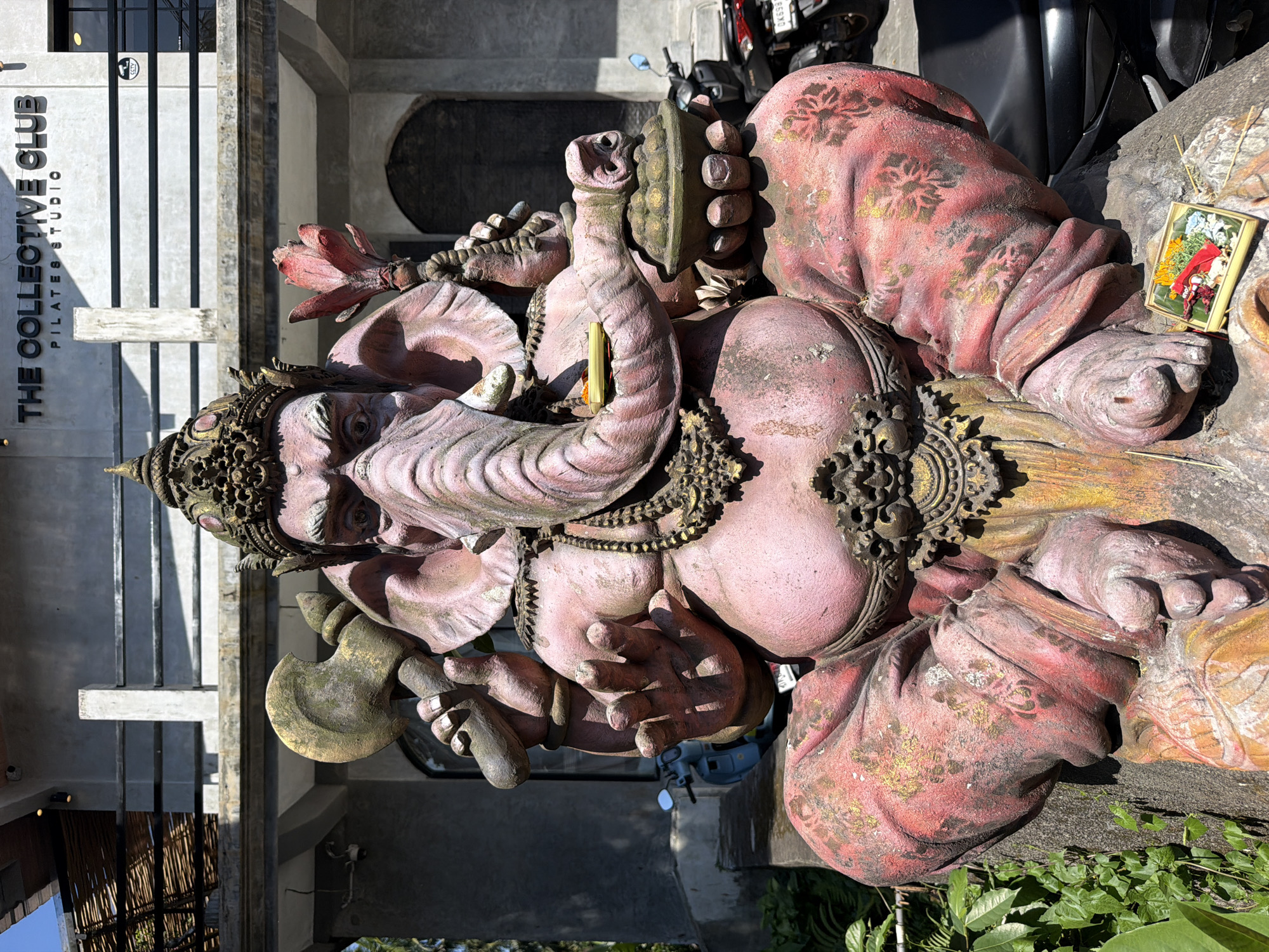 A large, pink Ganesha statue, adorned with a golden headdress and jewelry, is seated on a stone base. The statue depicts the elephant-headed deity with a large belly and is holding a battle axe in its right hand and a bowl of fruits in its left. A small offering of flowers and fruits is placed at the base of the statue. In the background, a building with the text "THE COLLECTIVE CLUB PILATES STUDIO" is visible, along with a blue scooter and a black motorcycle. The scene is outdoors, likely in a public space in Ubud District, Indonesia, given the religious iconography and architectural style. The lighting suggests it is daytime, with the sun casting shadows.
