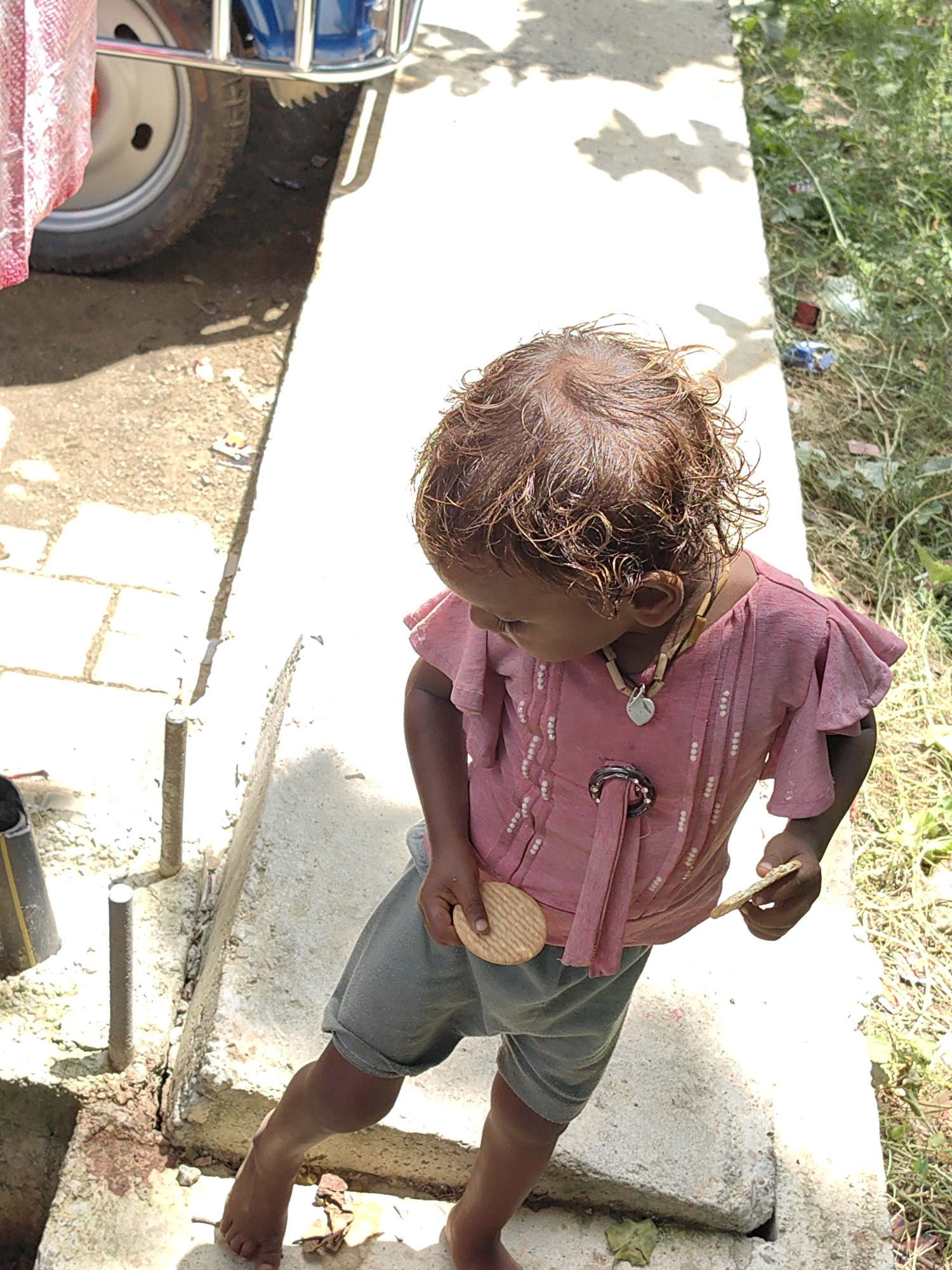 In Lumbini Bazaar, Nepal, a young child stands on a concrete ledge, dressed in a pink ruffled shirt and gray shorts. The child is holding a biscuit in each hand. The background shows dry soil, some green grass, and a portion of a blue vehicle with a red fabric draped over it. The scene appears to be outdoors on a sunny day.