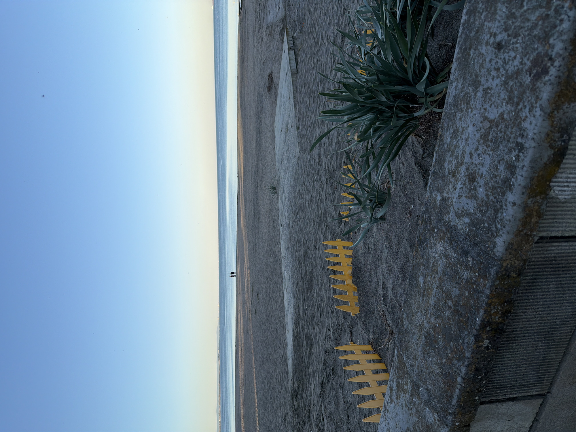 A slightly angled, landscape-oriented image depicts a beach scene, with the horizon line near the upper left. The sky transitions from light blue at the top to a yellowish hue near the horizon. The ocean stretches from the horizon to the middle ground, a strip of dark sand with two human figures in the distance. In the foreground, starting from the bottom right corner, is a weathered concrete structure and a patch of spiky green plants. Embedded in the sand are several bright yellow fence posts. The location is Unknown, Unknown.